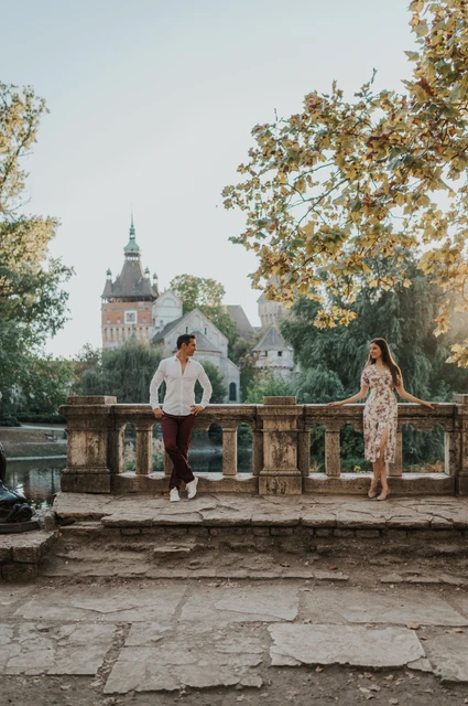 Castle rooftops beyond the balustrade at a Budapest couple photoshoot The couple standing apart beside a stone balustrade with Vajdahunyad Castle’s rooftops and leafy trees behind them during their couple photoshoot in Budapest.