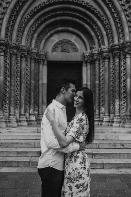 A castle portrait in black and white at a Budapest couple photoshoot Black-and-white portrait of the man kissing his smiling partner beside Vajdahunyad Castle’s carved doorway during their couple photoshoot in Budapest.