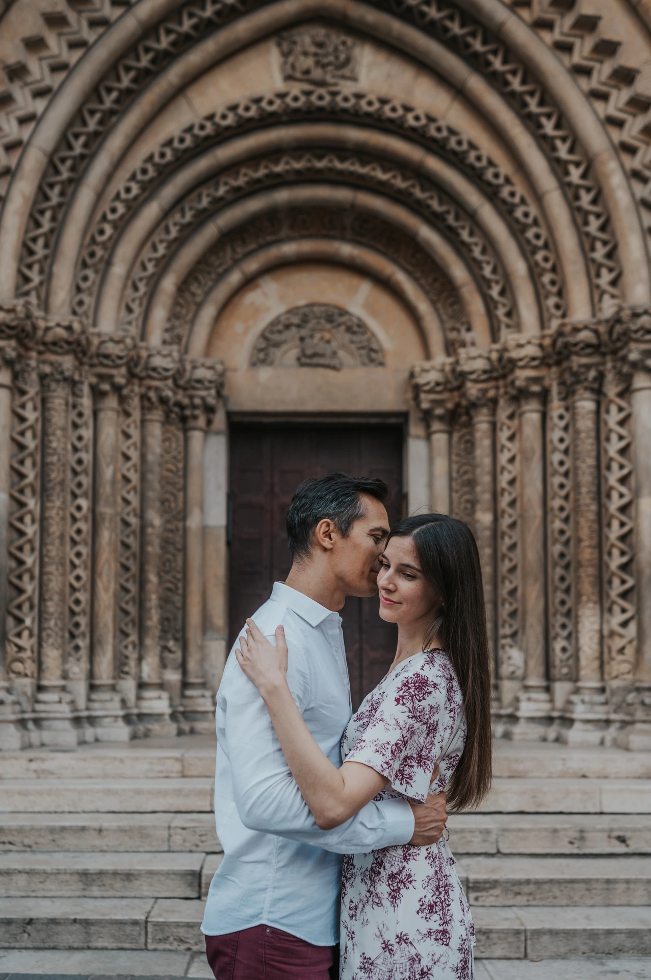 The man kissing his partner near her temple as she smiles beside Vajdahunyad Castle’s ornate doorway during their couple photoshoot in Budapest.