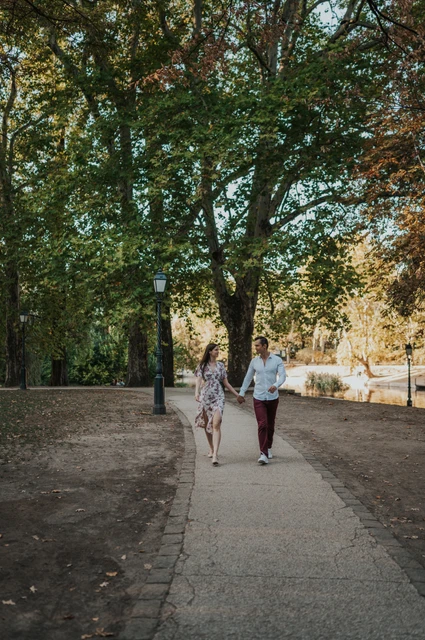 A leafy path beside the castle at a Budapest couple photoshoot A wide portrait of the couple walking hand in hand along a leafy path near Vajdahunyad Castle during their couple photoshoot in Budapest.