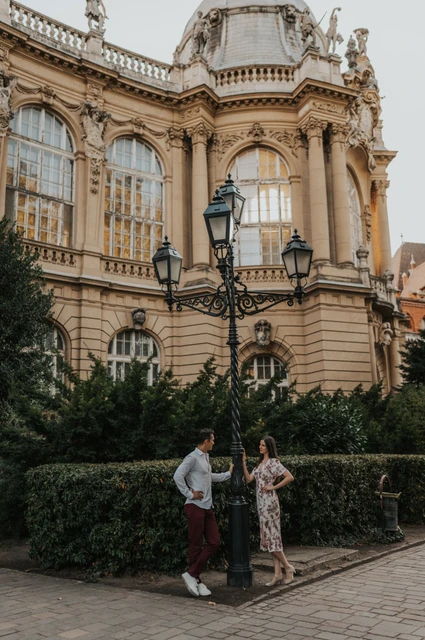 The couple beside an ornate black lamp and hedges beneath the pale palace façade at Vajdahunyad Castle during their couple photoshoot in Budapest.
