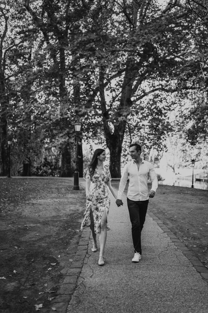 A park walk in black and white at a Budapest couple photoshoot Black-and-white portrait of the couple walking hand in hand on a tree-lined path near Vajdahunyad Castle during their couple photoshoot in Budapest.