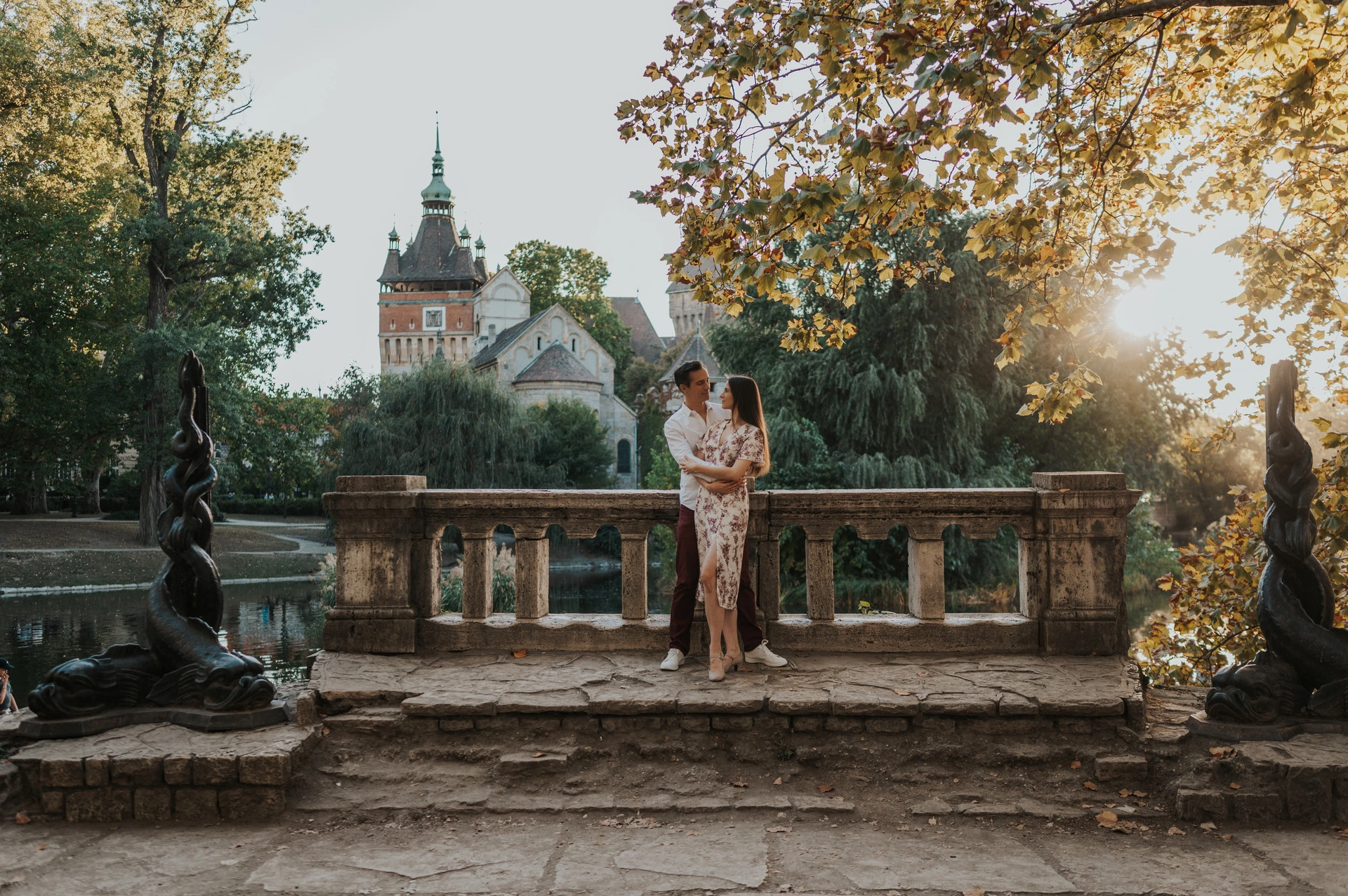 A wide view of the couple by a stone balustrade framed by a statue, golden leaves and Vajdahunyad Castle during their couple photoshoot in Budapest.