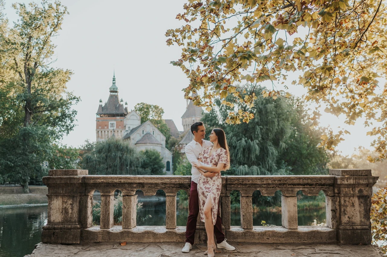 Sunlit leaves and a castle backdrop at a Budapest couple photoshoot A wide portrait of the couple embracing at a stone balustrade with Vajdahunyad Castle and sunlit leaves behind them during their couple photoshoot in Budapest.
