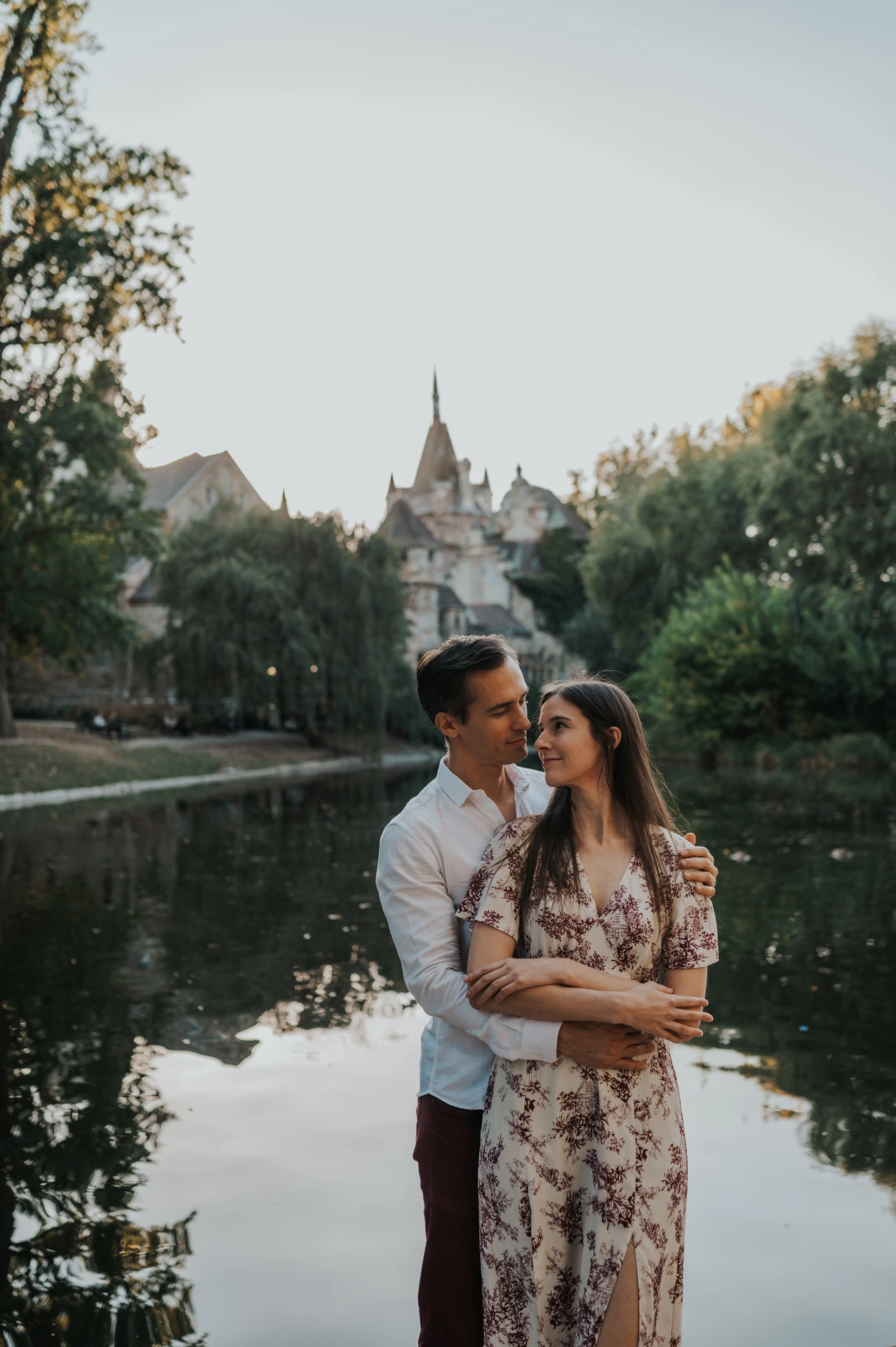 The man embracing his partner from behind beside the water with Vajdahunyad Castle and its reflection in the background during their couple photoshoot in Budapest.
