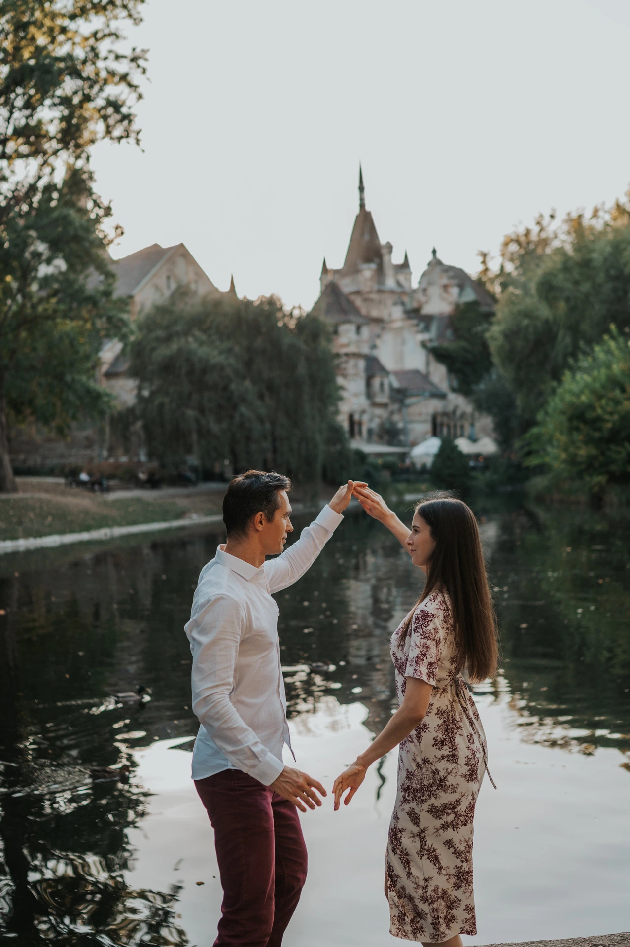 The couple raising their joined hands for a playful turn beside the water at Vajdahunyad Castle during their couple photoshoot in Budapest.