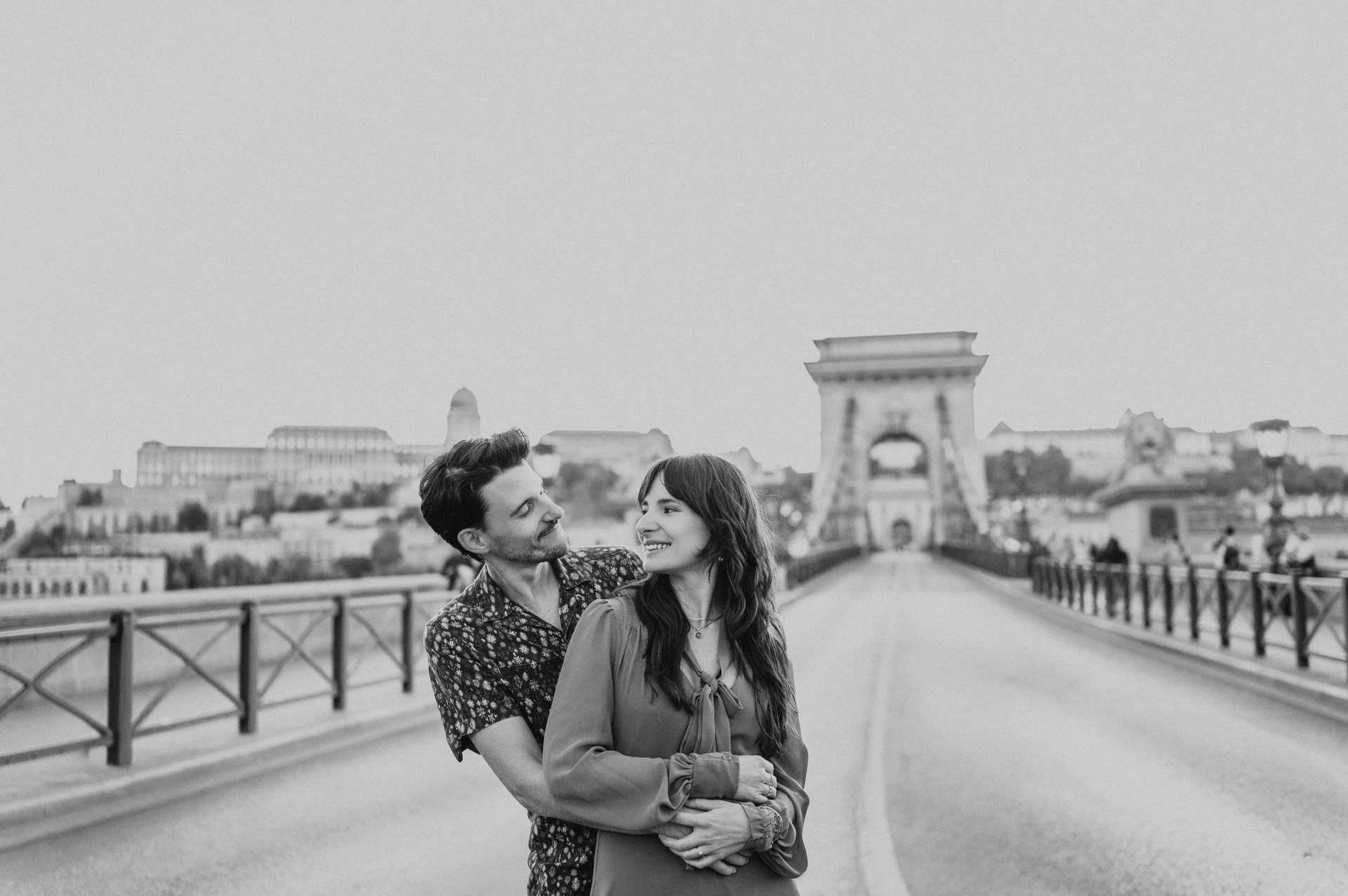Black-and-white landscape portrait of the couple embracing and smiling at one another on Chain Bridge during their Budapest engagement photoshoot.