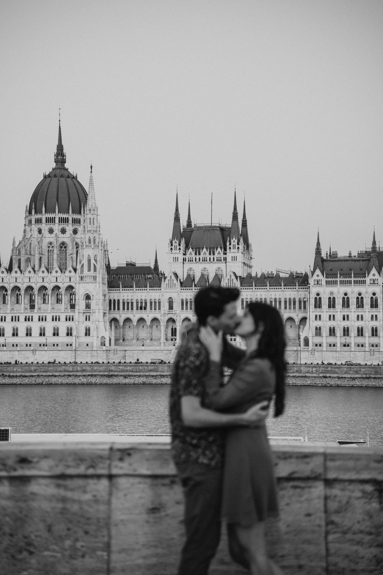 Budapest engagement photoshoot: A black-and-white kiss across from Parliament Black-and-white photograph of the couple kissing beside the stone wall, with Parliament across the river behind them during their Budapest engagement photoshoot.