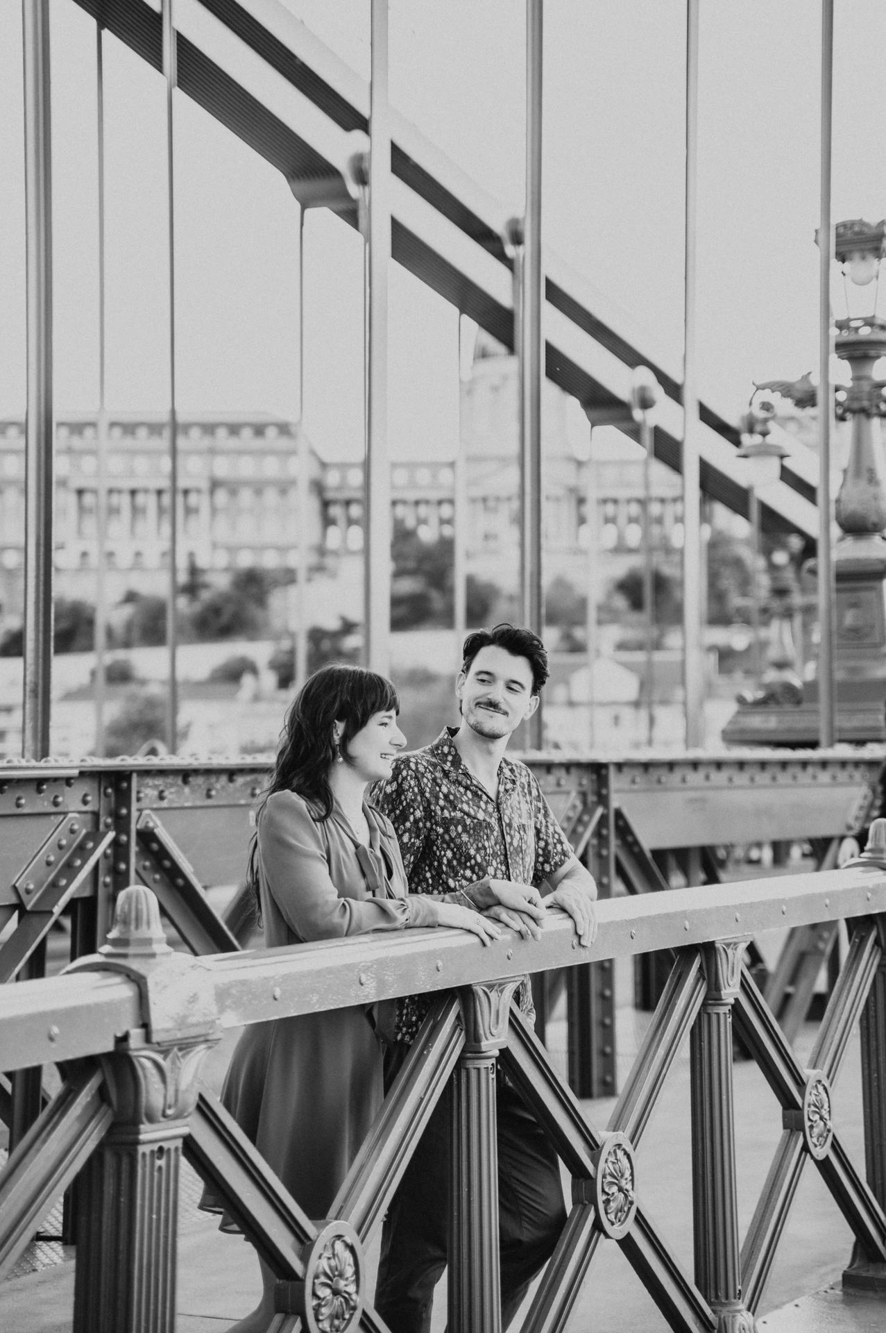 Budapest engagement photoshoot: A black-and-white portrait at the railing Black-and-white view of the couple sharing a smile beside Chain Bridge’s metal railing and suspension rods during their Budapest engagement photoshoot.