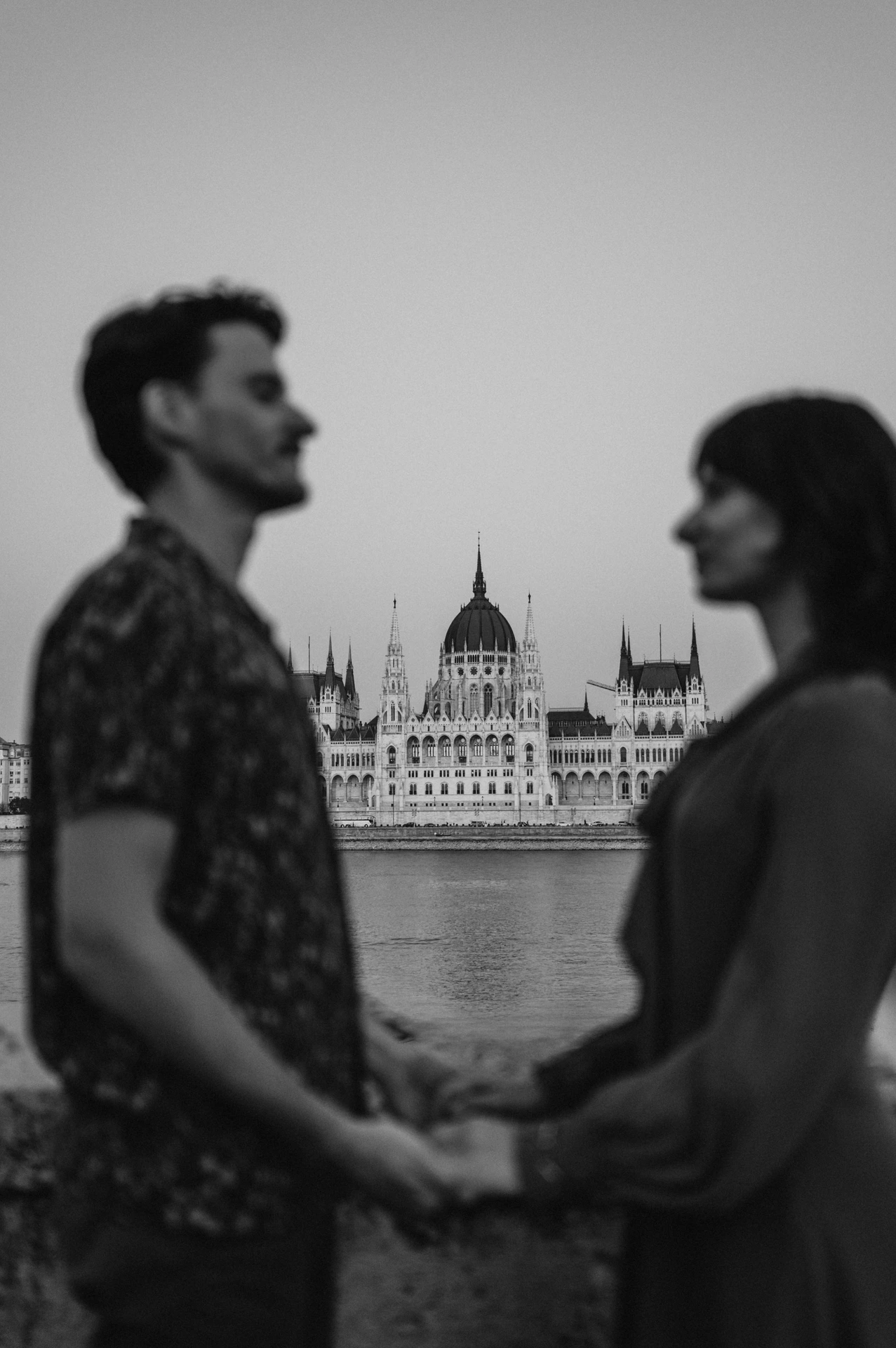 Budapest engagement photoshoot: A black-and-white portrait holding hands Black-and-white photograph of the couple holding hands and looking at each other, with Parliament across the river behind them during their Budapest engagement photoshoot.
