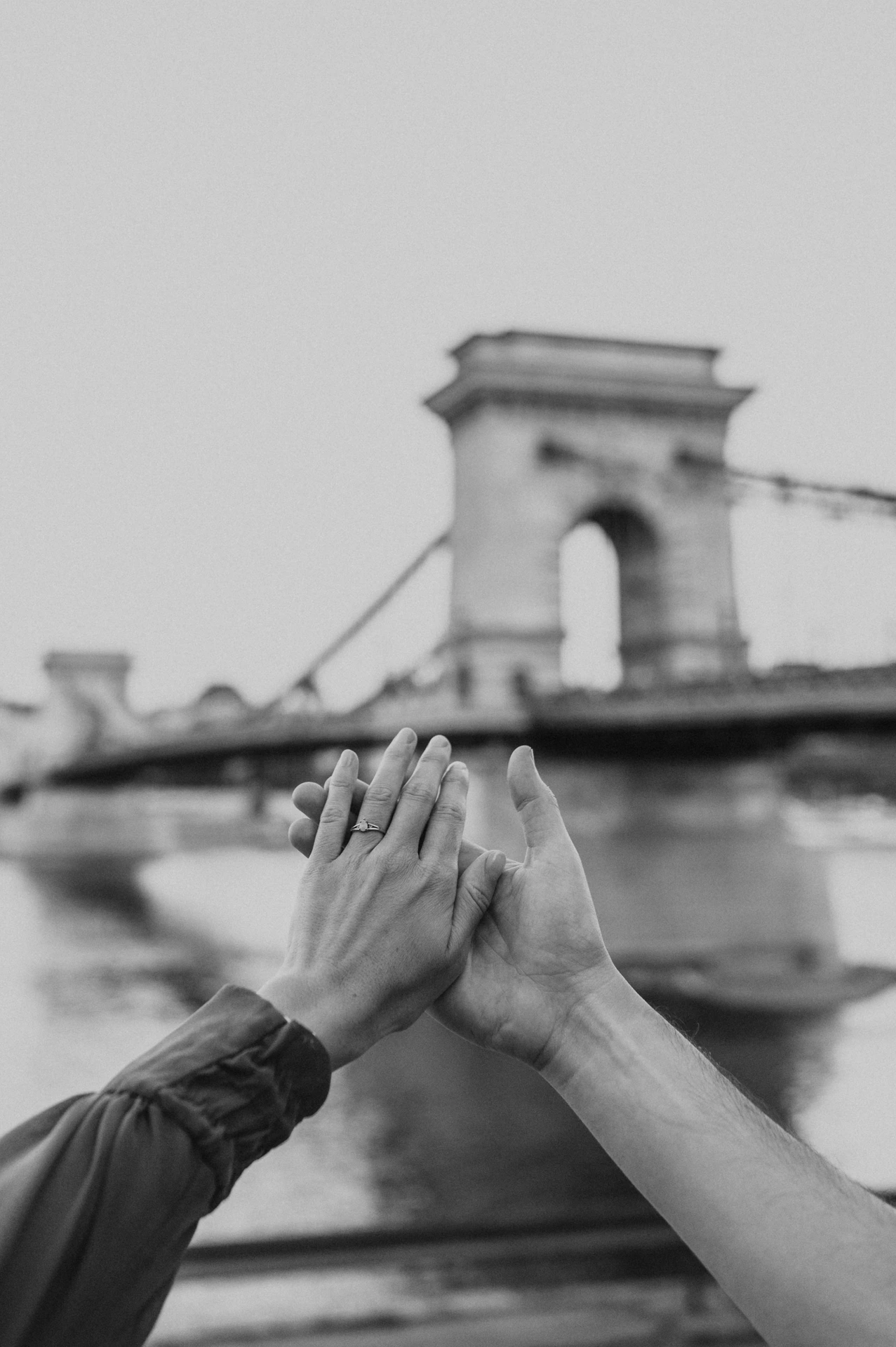 Budapest engagement photoshoot: A black-and-white ring detail by the bridge Black-and-white close-up of the couple’s hands together and the engagement ring, with Chain Bridge softly behind them during their Budapest engagement photoshoot.