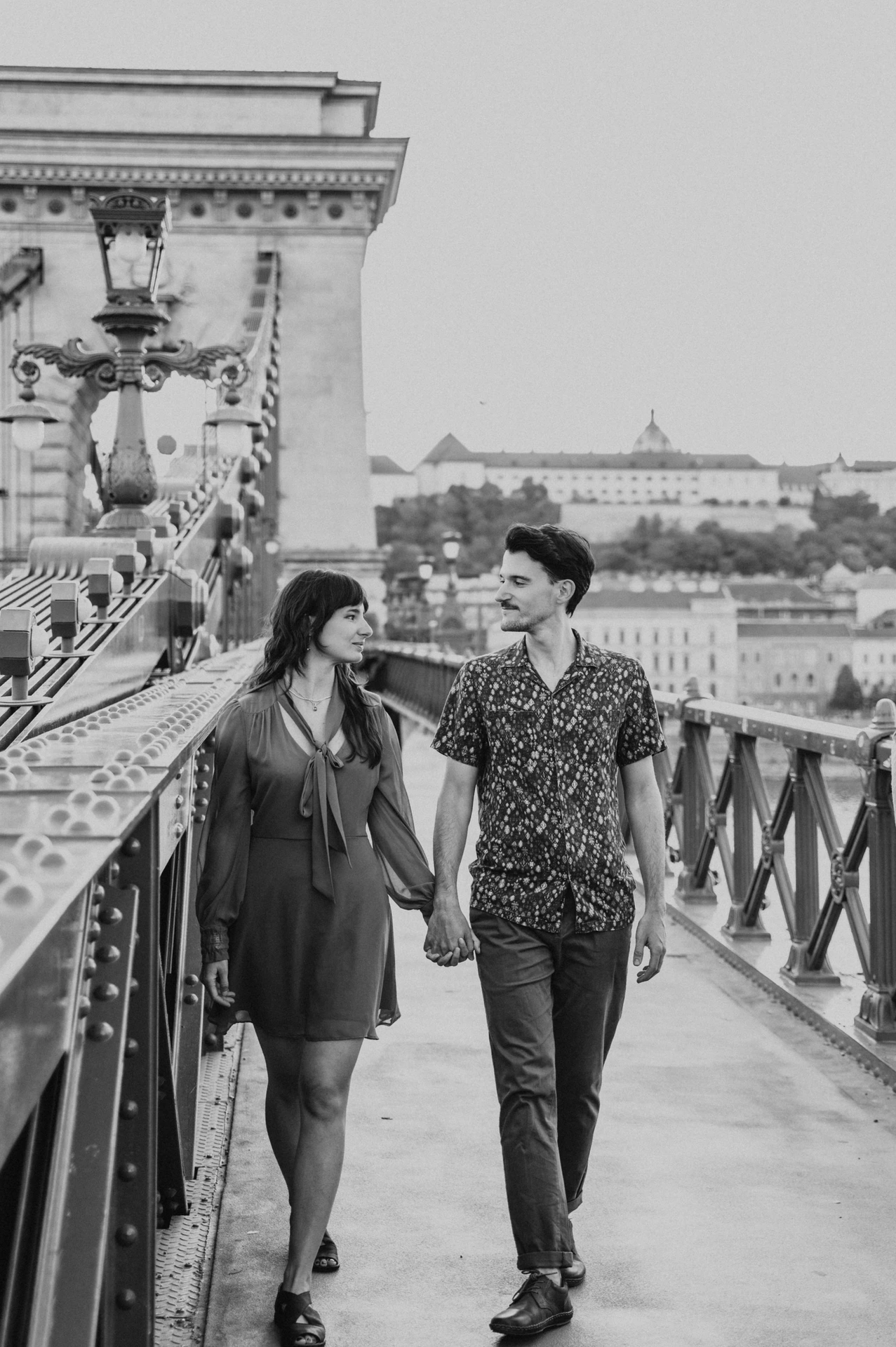 Budapest engagement photoshoot: A black-and-white stroll past the bridge lamps Black-and-white photograph of the couple holding hands on Chain Bridge, with its lamps, railing and stone tower around them during their Budapest engagement photoshoot.