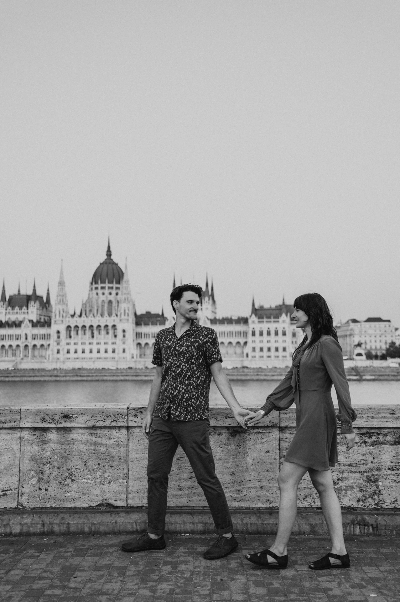 Budapest engagement photoshoot: A black-and-white walk beside the Danube Black-and-white photograph of the couple walking together by the river wall, with Parliament’s dome across the water during their Budapest engagement photoshoot.