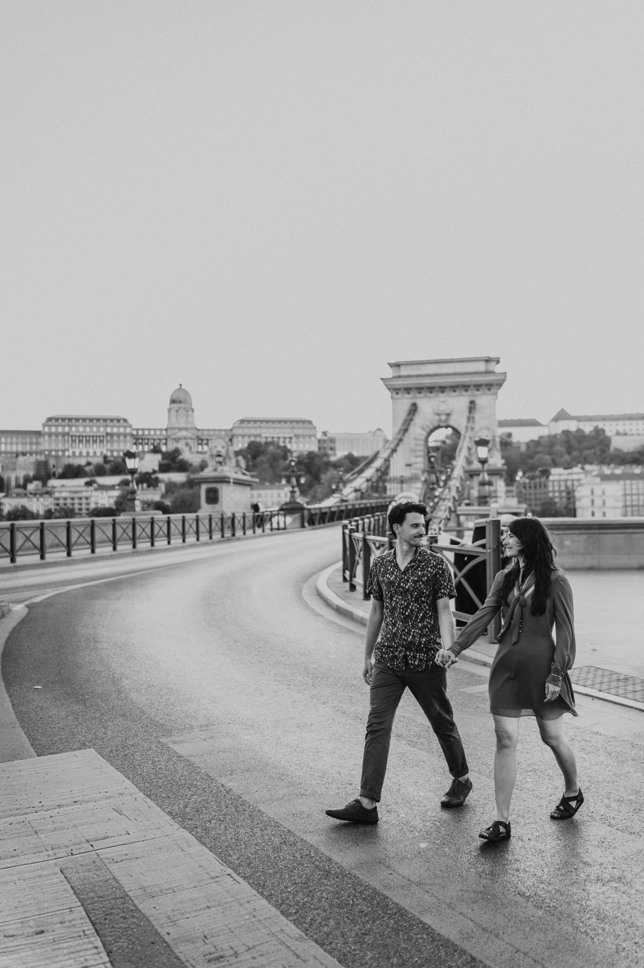 Black-and-white view of the couple walking together along the bridge, with the curved walkway and stone tower behind them during their Budapest engagement photoshoot.