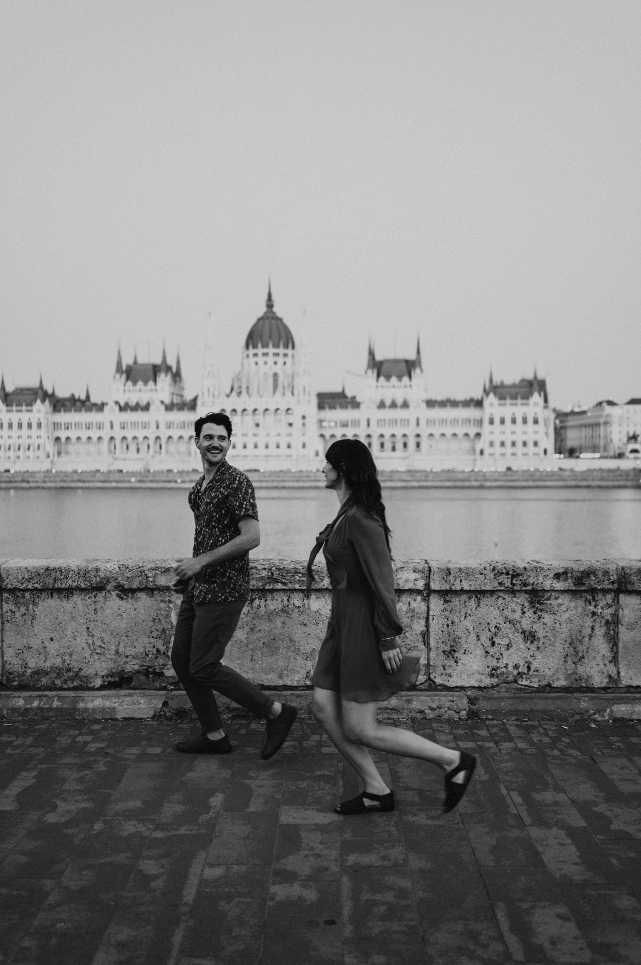Budapest engagement photoshoot: A black-and-white walk with Parliament in the backdrop Black-and-white photograph of the couple walking hand in hand along the riverside, with Parliament across the water during their Budapest engagement photoshoot.