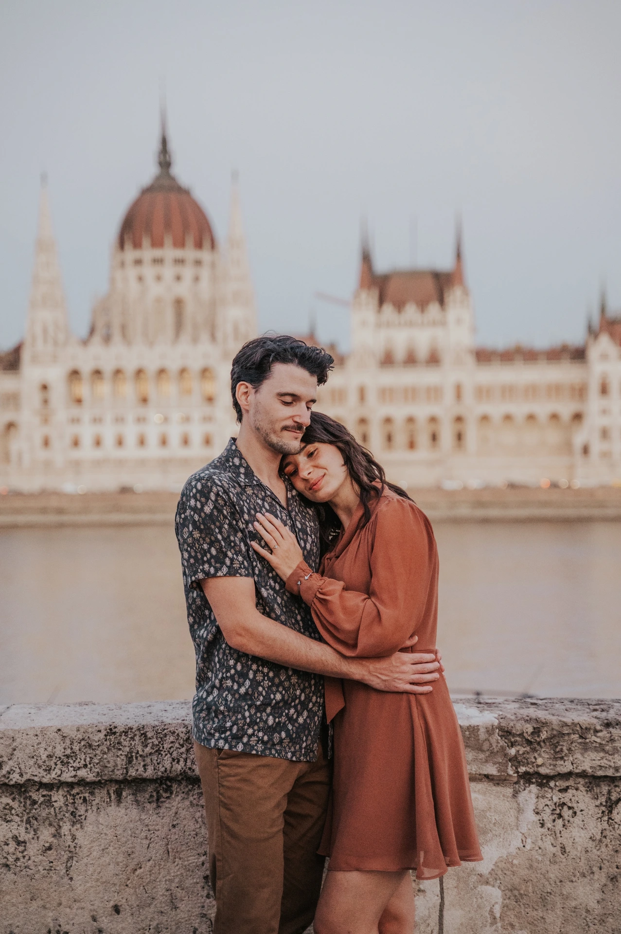 Budapest engagement photoshoot: A close kiss beside the Danube A close portrait of the couple cuddling by the river wall, with a kiss on the cheek and Parliament softly behind them during their Budapest engagement photoshoot.