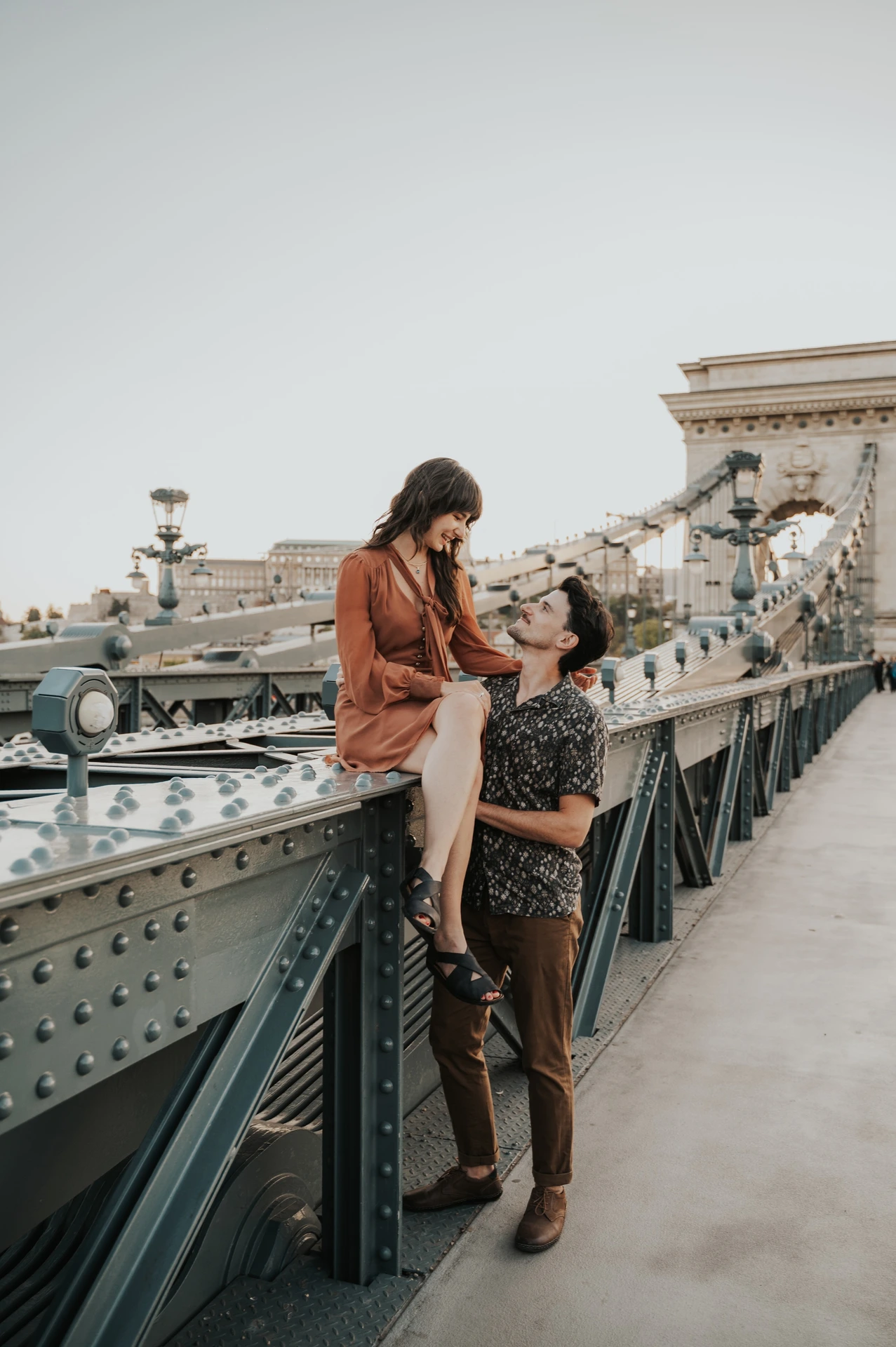 Budapest engagement photoshoot: A closer portrait along the bridge rail A closer view of the couple embracing beside Chain Bridge’s dark metal railing and decorative street lamps during their Budapest engagement photoshoot.