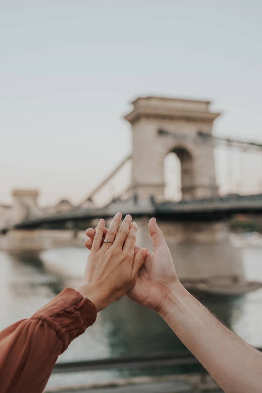 Budapest engagement photoshoot: A closer ring detail beside Chain Bridge A vertical close-up of the couple’s hands and engagement ring, with Chain Bridge blurred in the riverside background during their Budapest engagement photoshoot.