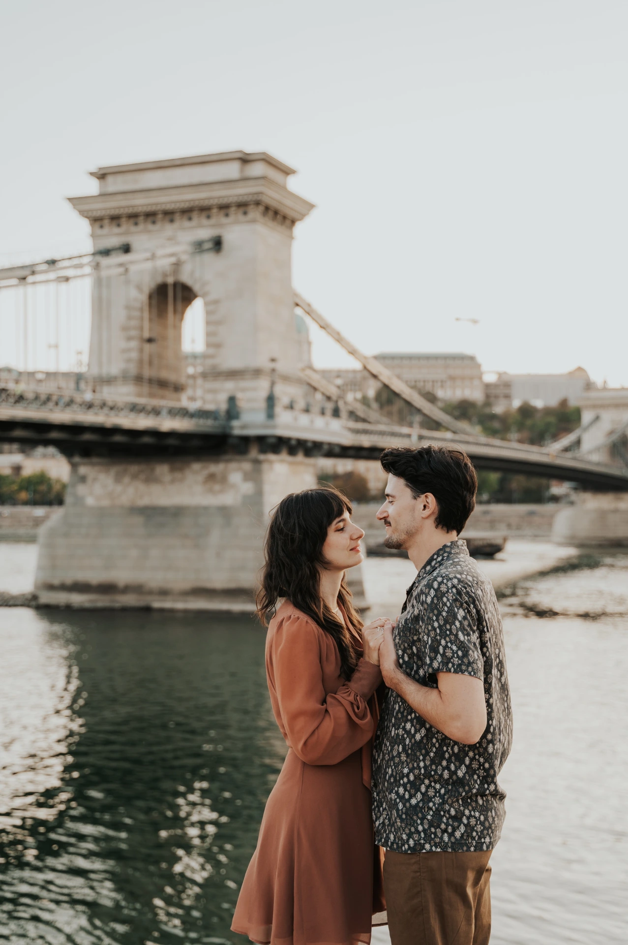 Budapest engagement photoshoot: A closer riverside portrait by the bridge A closer vertical view of the couple holding hands beside the river, framed by Chain Bridge’s pale stone tower and suspension chains during their Budapest engagement photoshoot.