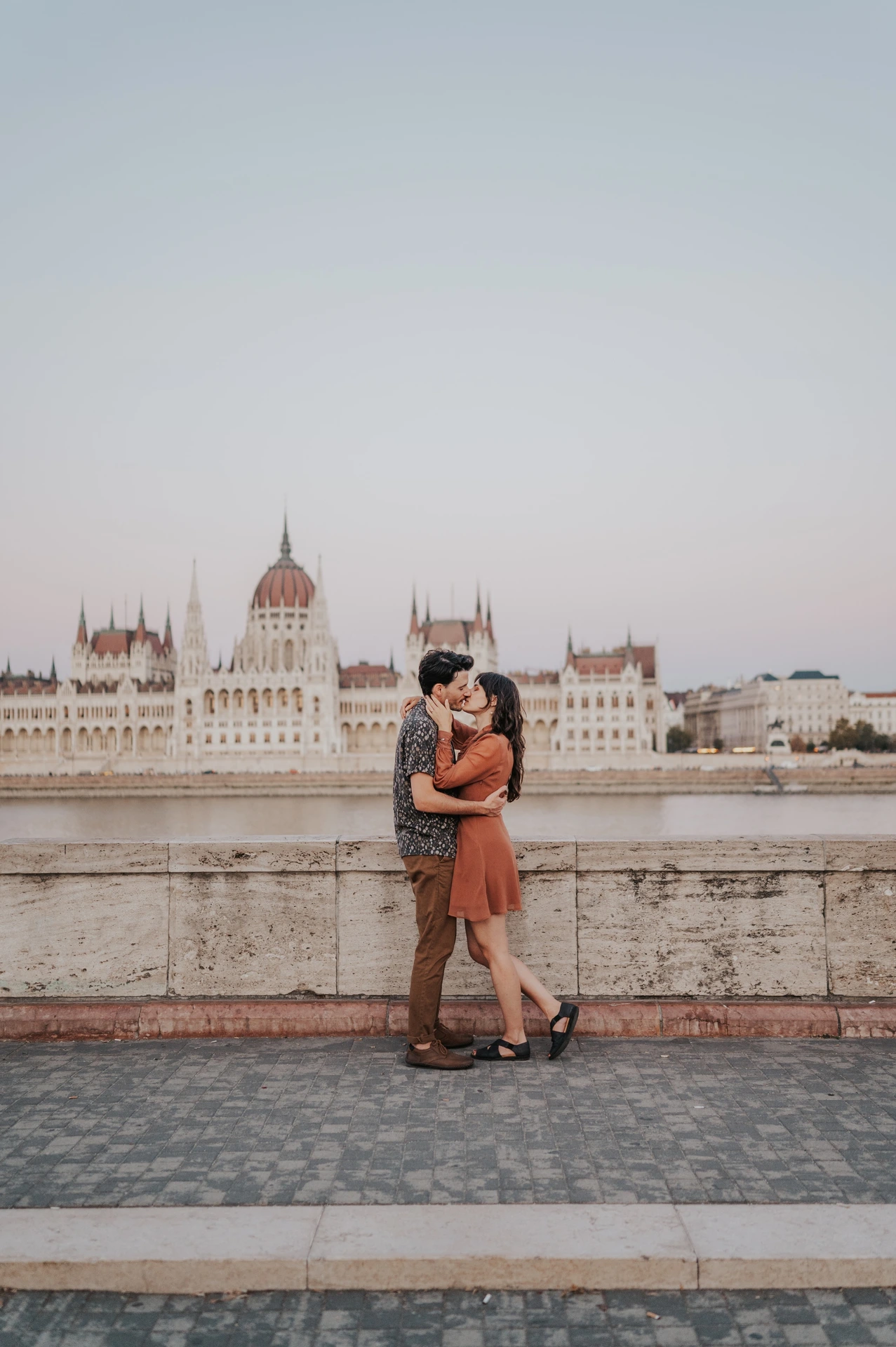 Budapest engagement photoshoot: A full-length embrace beside the Danube A full-length portrait of the couple embracing beside the riverside stone wall, with Parliament and the evening sky across the water during their Budapest engagement photoshoot.