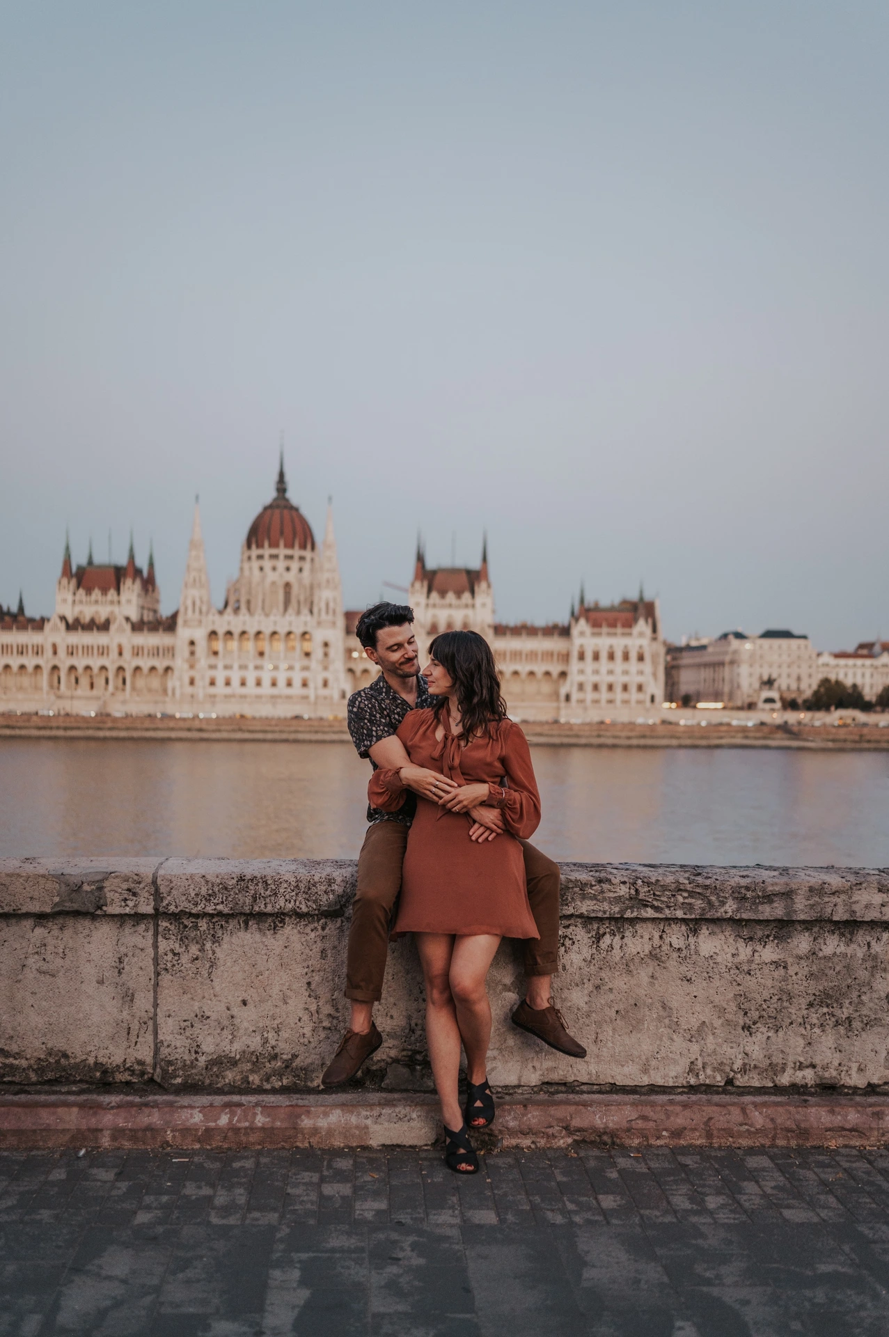 A full-length view of the couple close together beside the river wall, with Parliament’s dome and façade across the Danube during their Budapest engagement photoshoot.