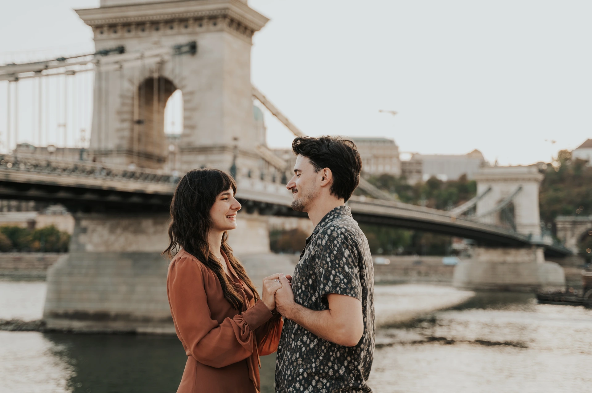 A closer landscape photograph of the couple holding hands beside the Danube, with Chain Bridge’s stone tower behind them during their Budapest engagement photoshoot.