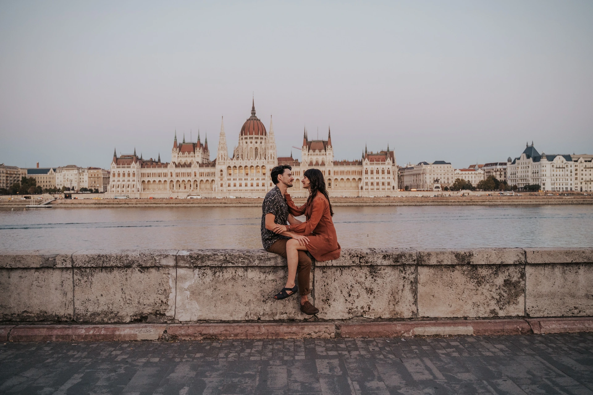 Budapest engagement photoshoot: A landscape portrait of the couple and Parliament A landscape photograph of the couple seated together on the river wall, with Parliament’s full façade and dome across the Danube during their Budapest engagement photoshoot.