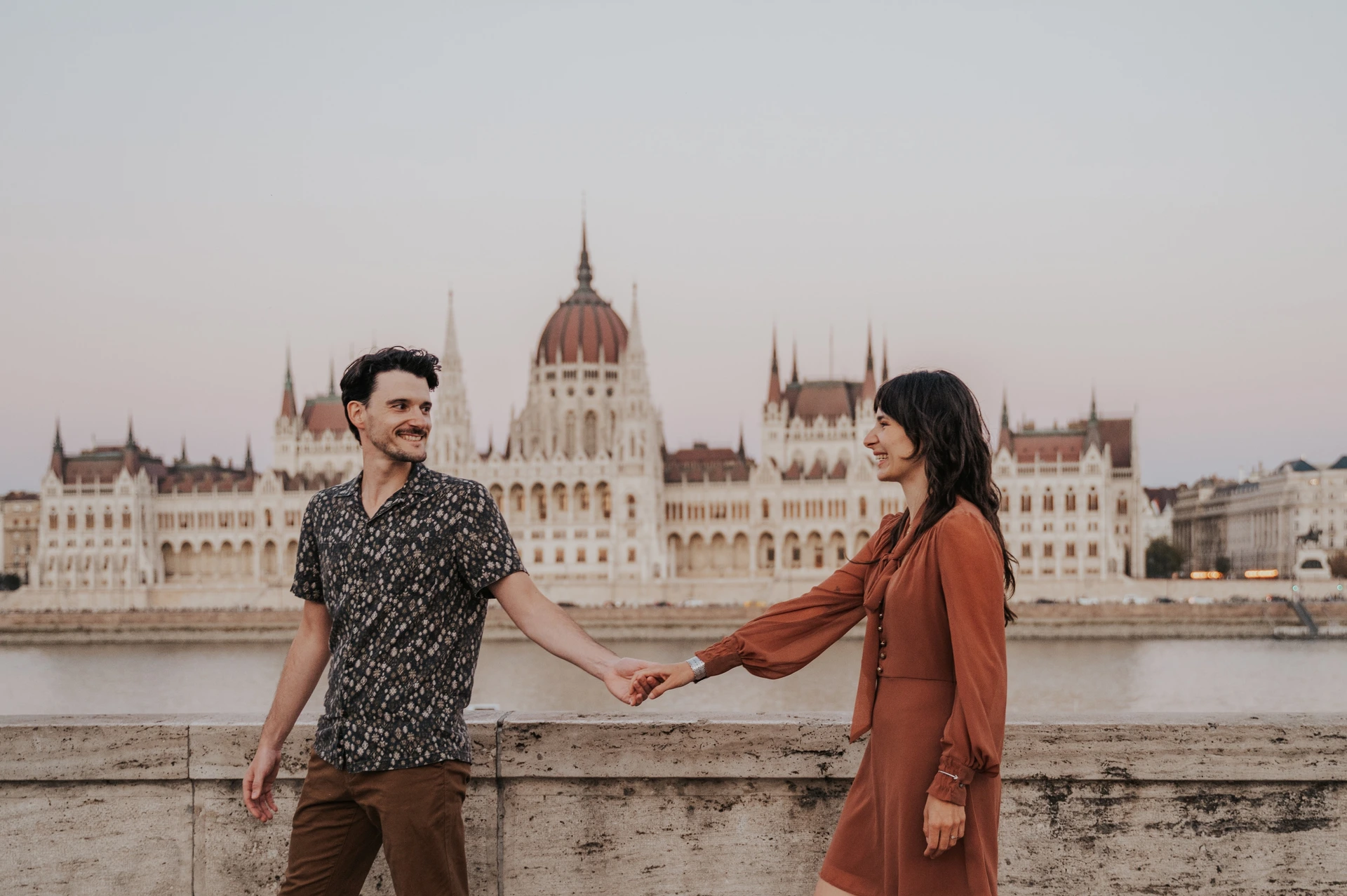 Budapest engagement photoshoot: A landscape walk with Parliament in the backdrop A landscape photograph of the couple smiling at one another while holding hands, with Parliament’s dome and façade across the river during their Budapest engagement photoshoot.