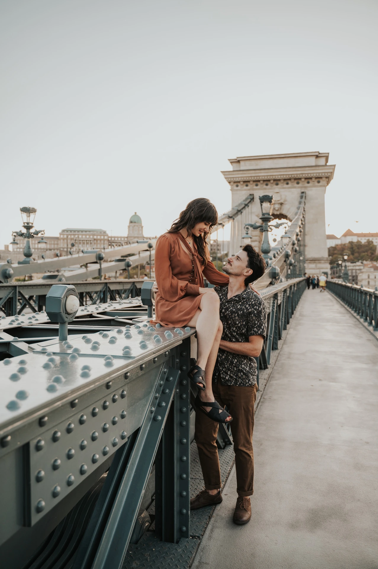 The couple embracing beside Chain Bridge’s metal railing, with one partner perched on the rail and the stone tower behind them during their Budapest engagement photoshoot.