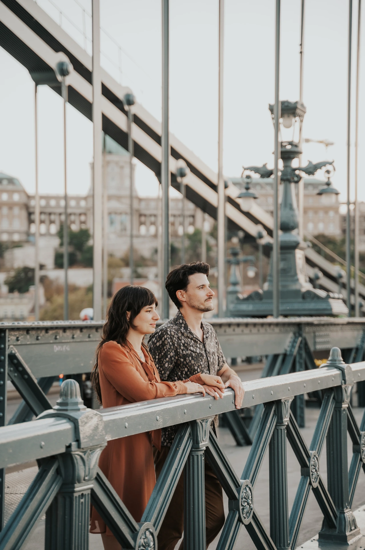Budapest engagement photoshoot: A quiet moment above the Danube The couple resting together against Chain Bridge’s metal railing, with the Danube and city buildings beyond them during their Budapest engagement photoshoot.
