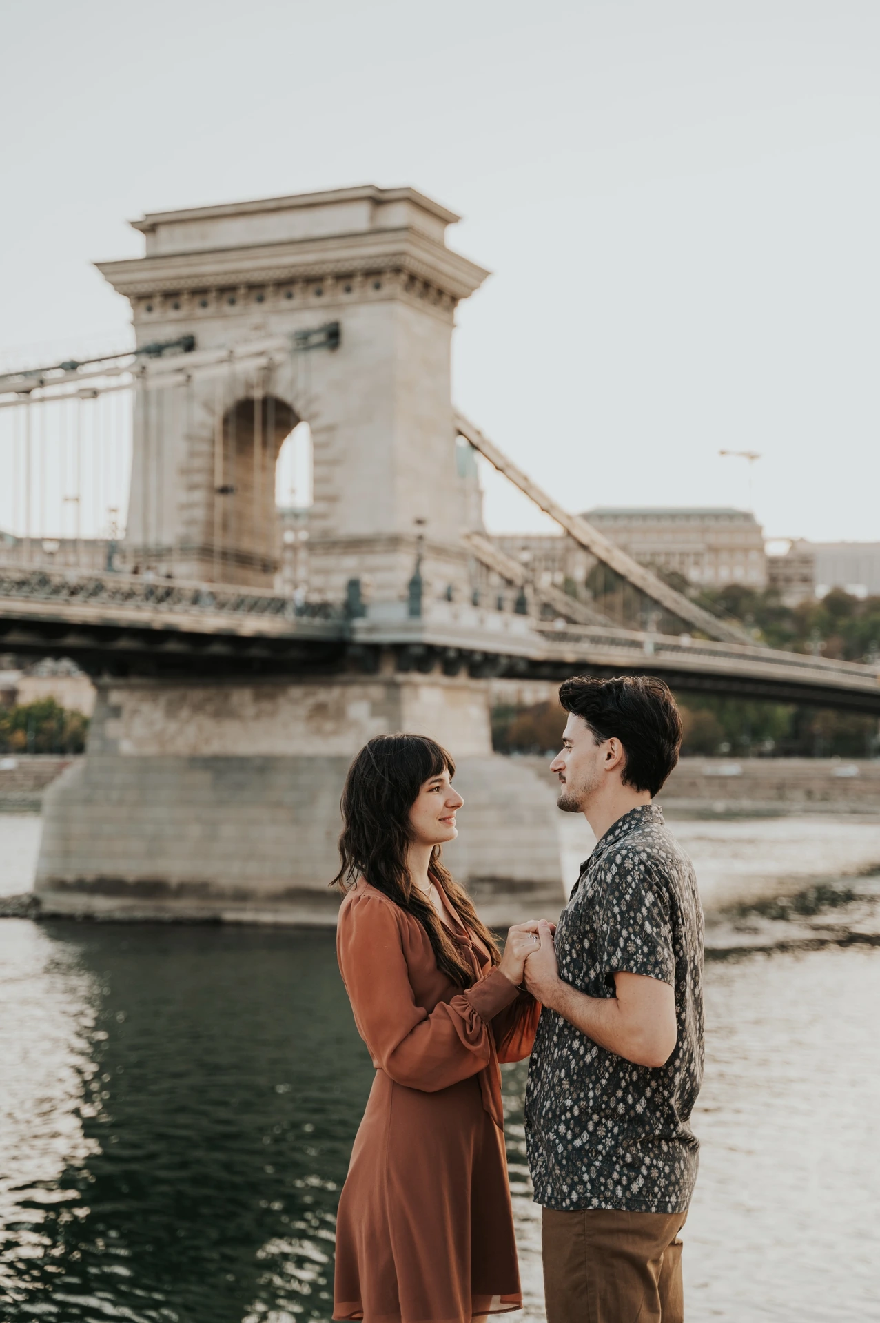 Budapest engagement photoshoot: A quiet smile with Chain Bridge in the backdrop The couple holding hands and smiling by the river, with Chain Bridge crossing the water behind them during their Budapest engagement photoshoot.