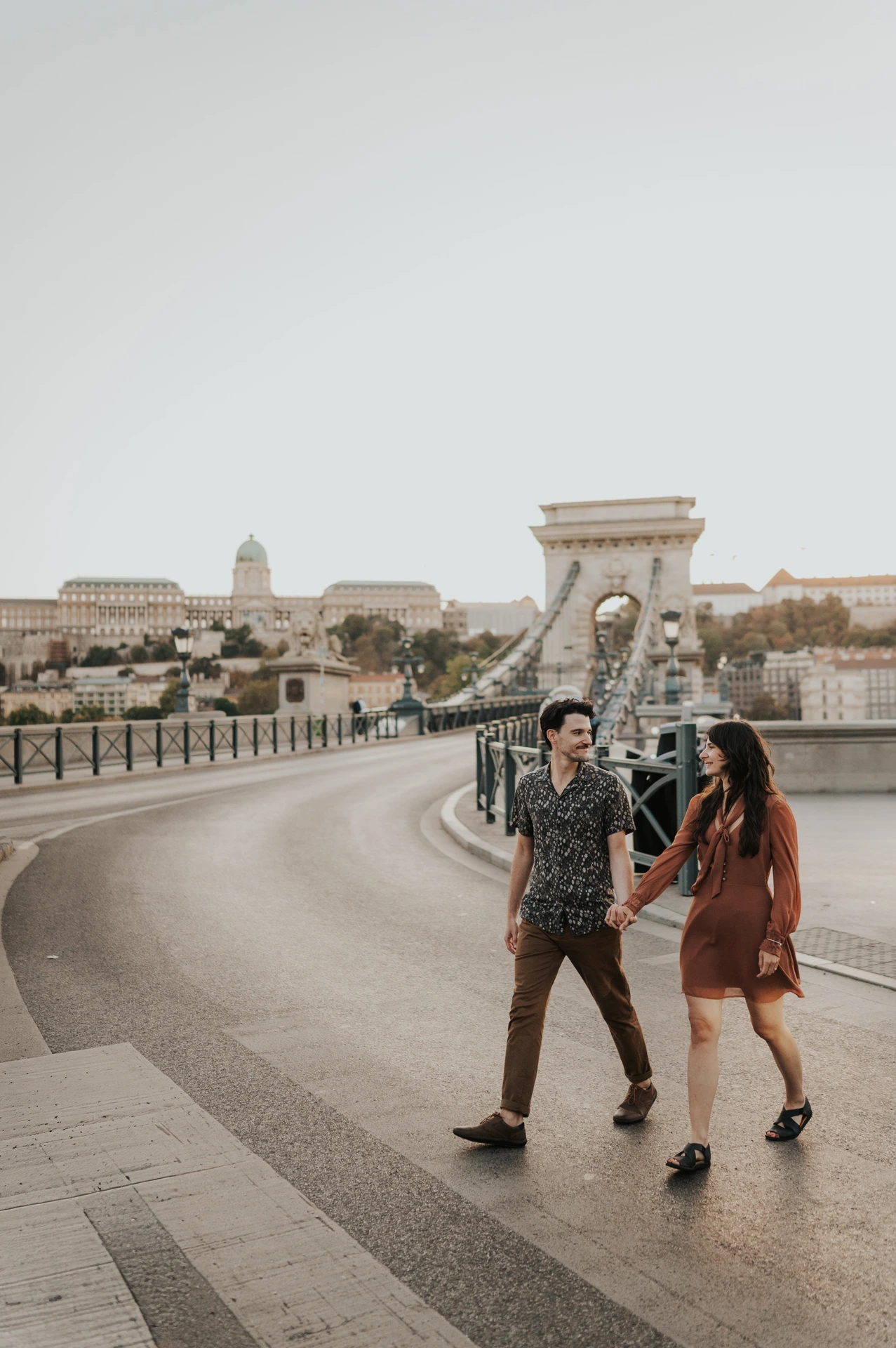 Budapest engagement photoshoot: A relaxed walk along the bridge The couple walking hand in hand along Chain Bridge, with its metal railings, street lamps and stone tower behind them during their Budapest engagement photoshoot.