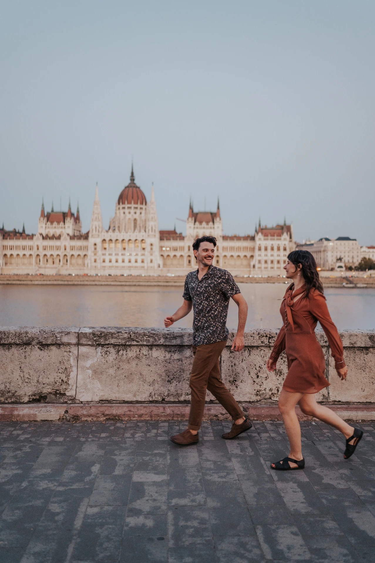 The couple walking hand in hand along the riverside paving, with Parliament stretching across the river behind them during their Budapest engagement photoshoot.