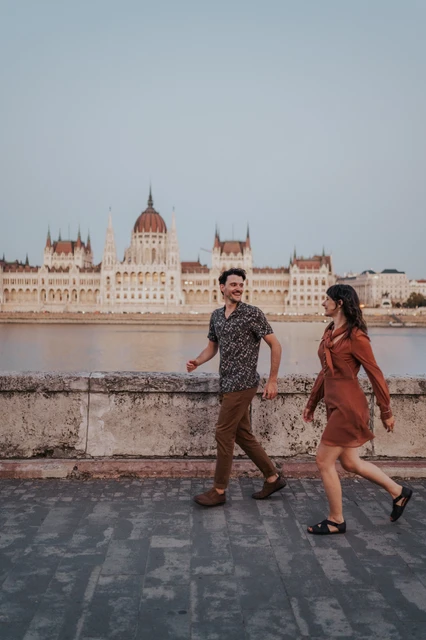 Budapest engagement photoshoot: A relaxed walk along the Danube The couple walking hand in hand along the riverside paving, with Parliament stretching across the river behind them during their Budapest engagement photoshoot.