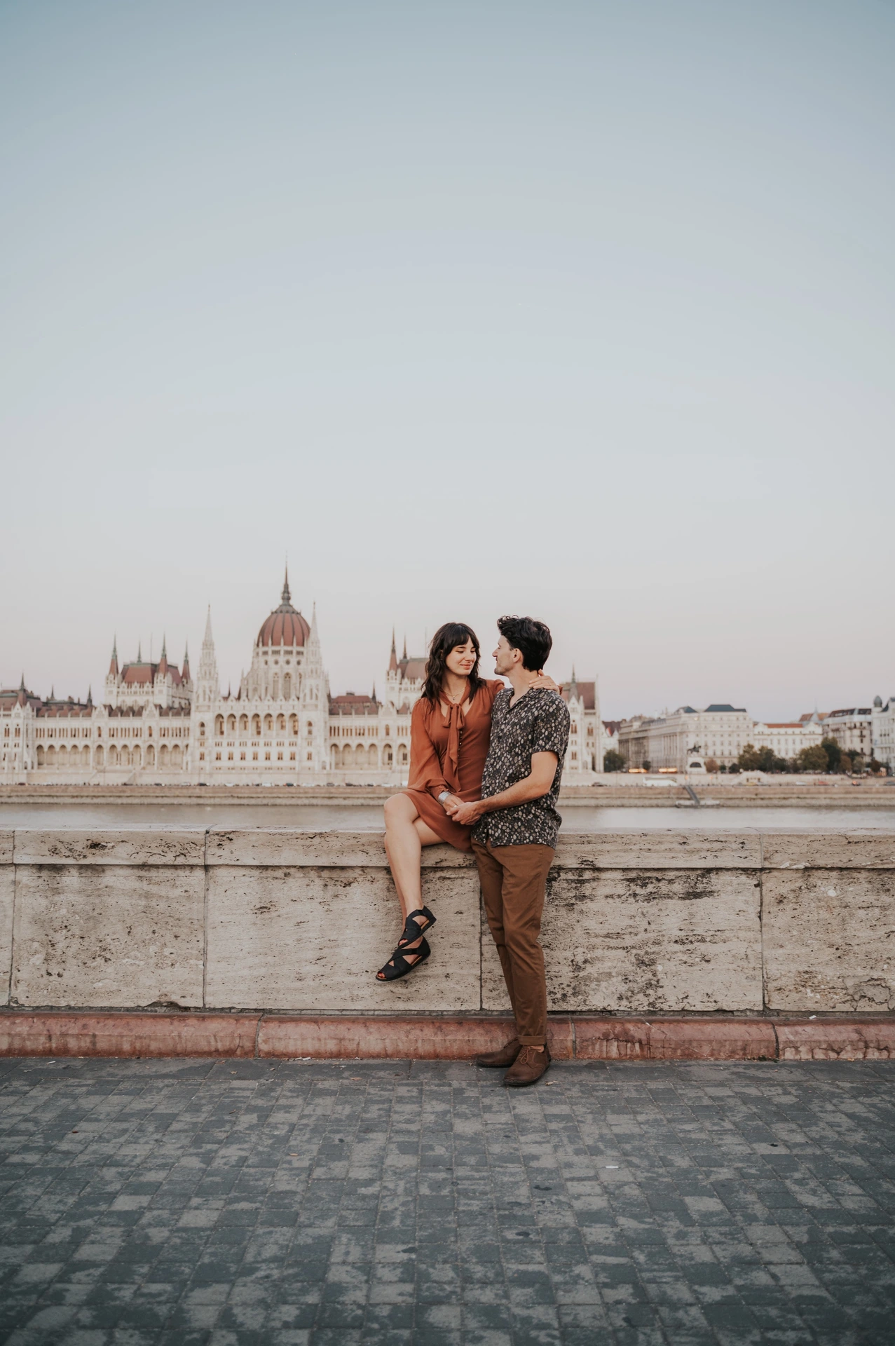 Budapest engagement photoshoot: A riverside kiss with Parliament across the water The couple kissing beside the riverside stone wall, with the Hungarian Parliament across the Danube behind them during their Budapest engagement photoshoot.