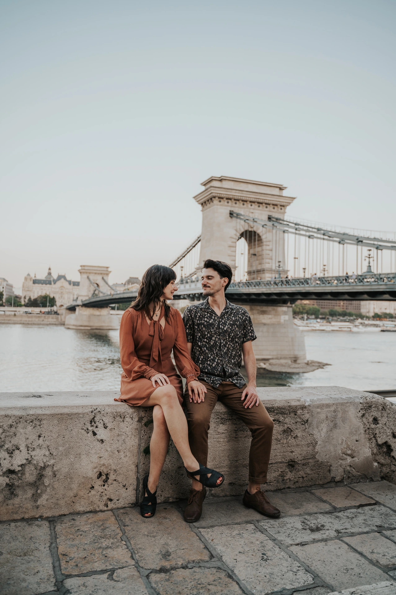 Budapest engagement photoshoot: A seated portrait with Chain Bridge behind them The couple sitting together on the riverside stone ledge, with Chain Bridge’s tower and suspension chains crossing the Danube behind them during their Budapest engagement photoshoot.