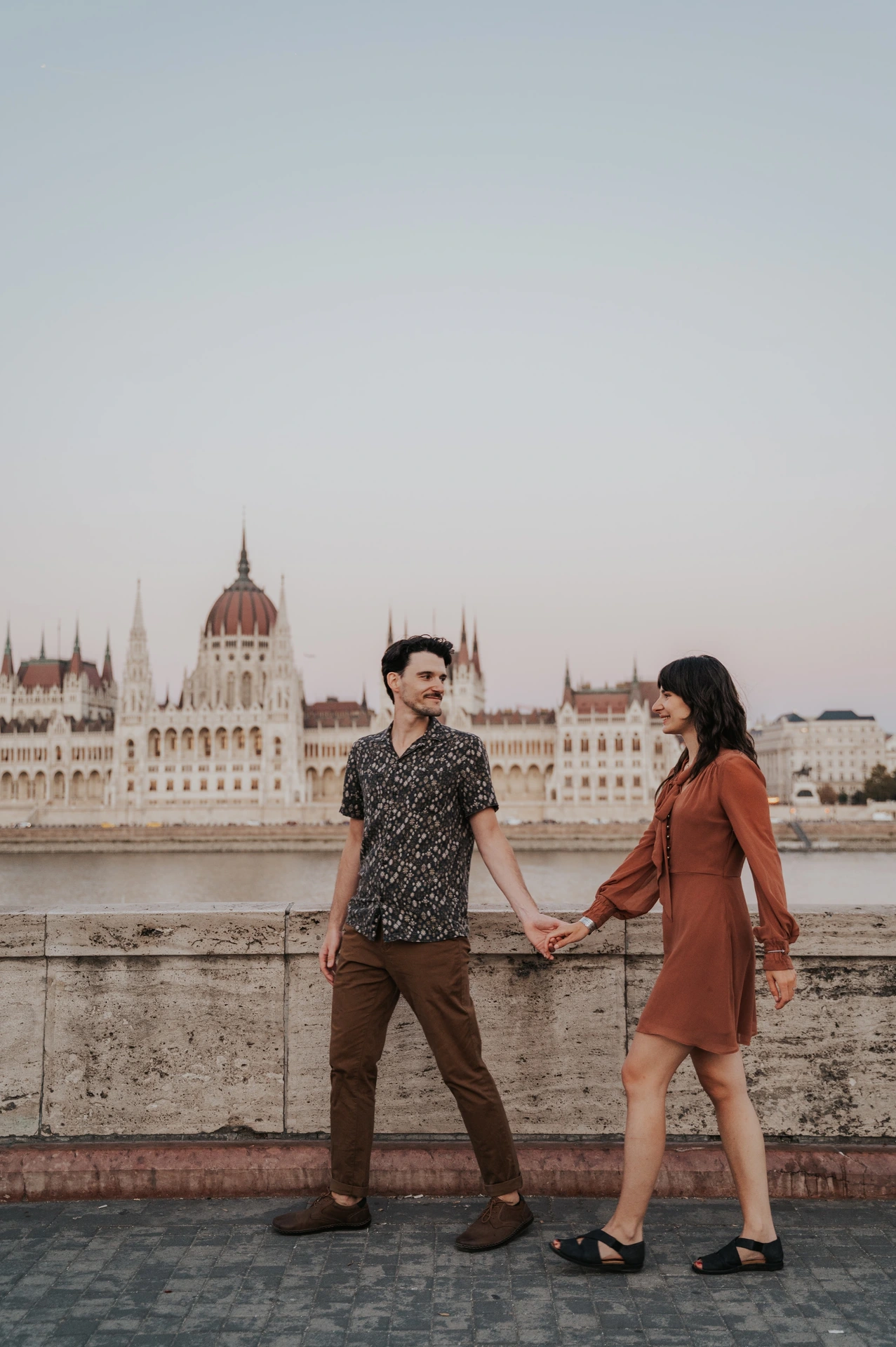 The couple looking towards one another while walking hand in hand along the riverside, with Parliament behind them during their Budapest engagement photoshoot.