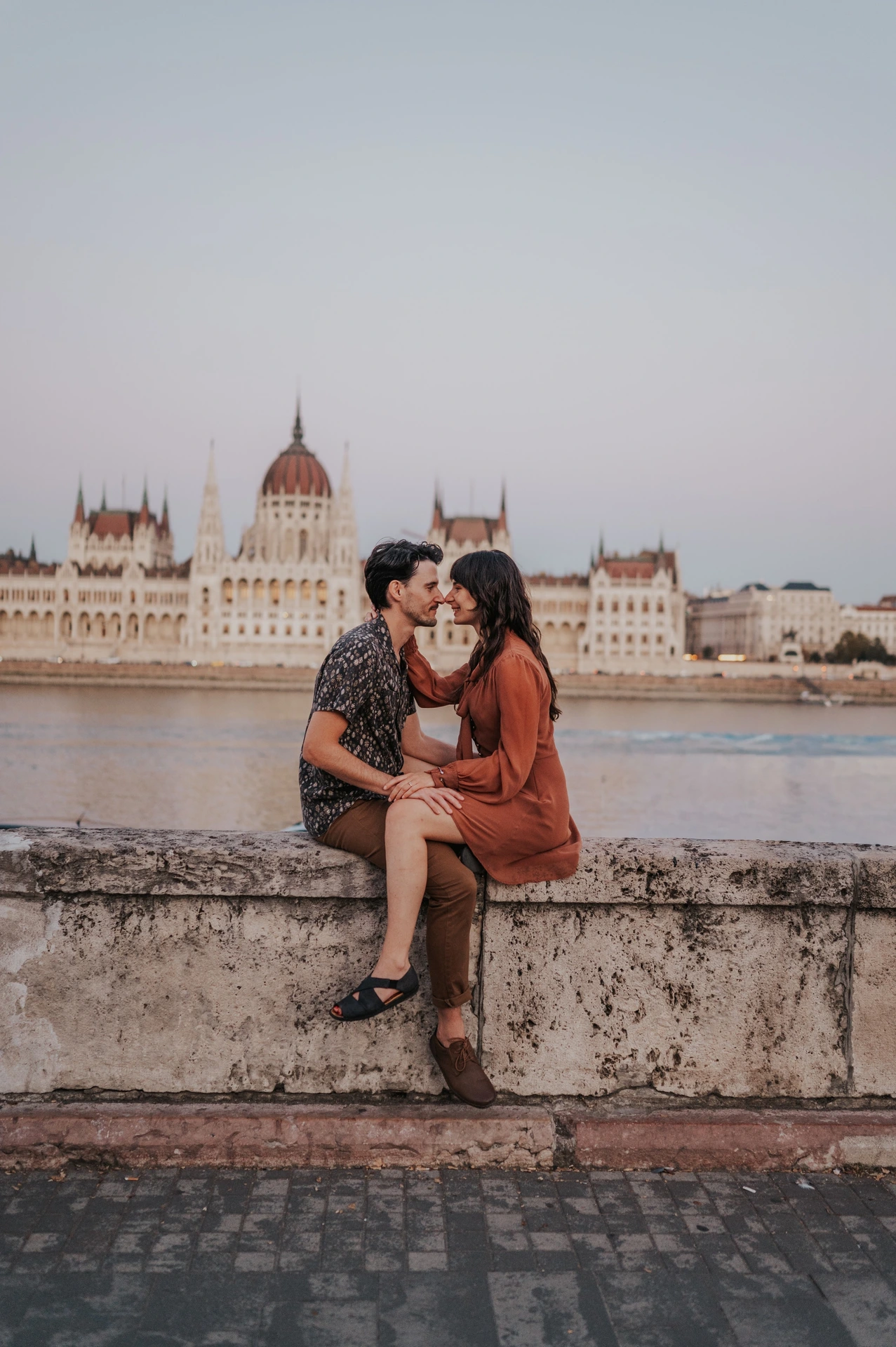 Budapest engagement photoshoot: A shared moment on the riverside ledge The couple looking towards one another while seated closely on the river wall, with Parliament across the water behind them during their Budapest engagement photoshoot.