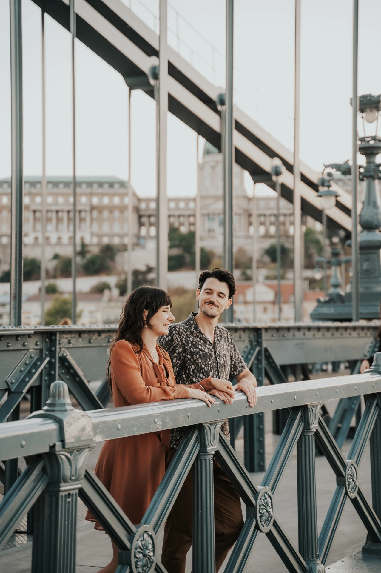 Budapest engagement photoshoot: A shared smile against the bridge rail The couple smiling together as they lean against Chain Bridge’s railing, with its vertical suspension rods behind them during their Budapest engagement photoshoot.
