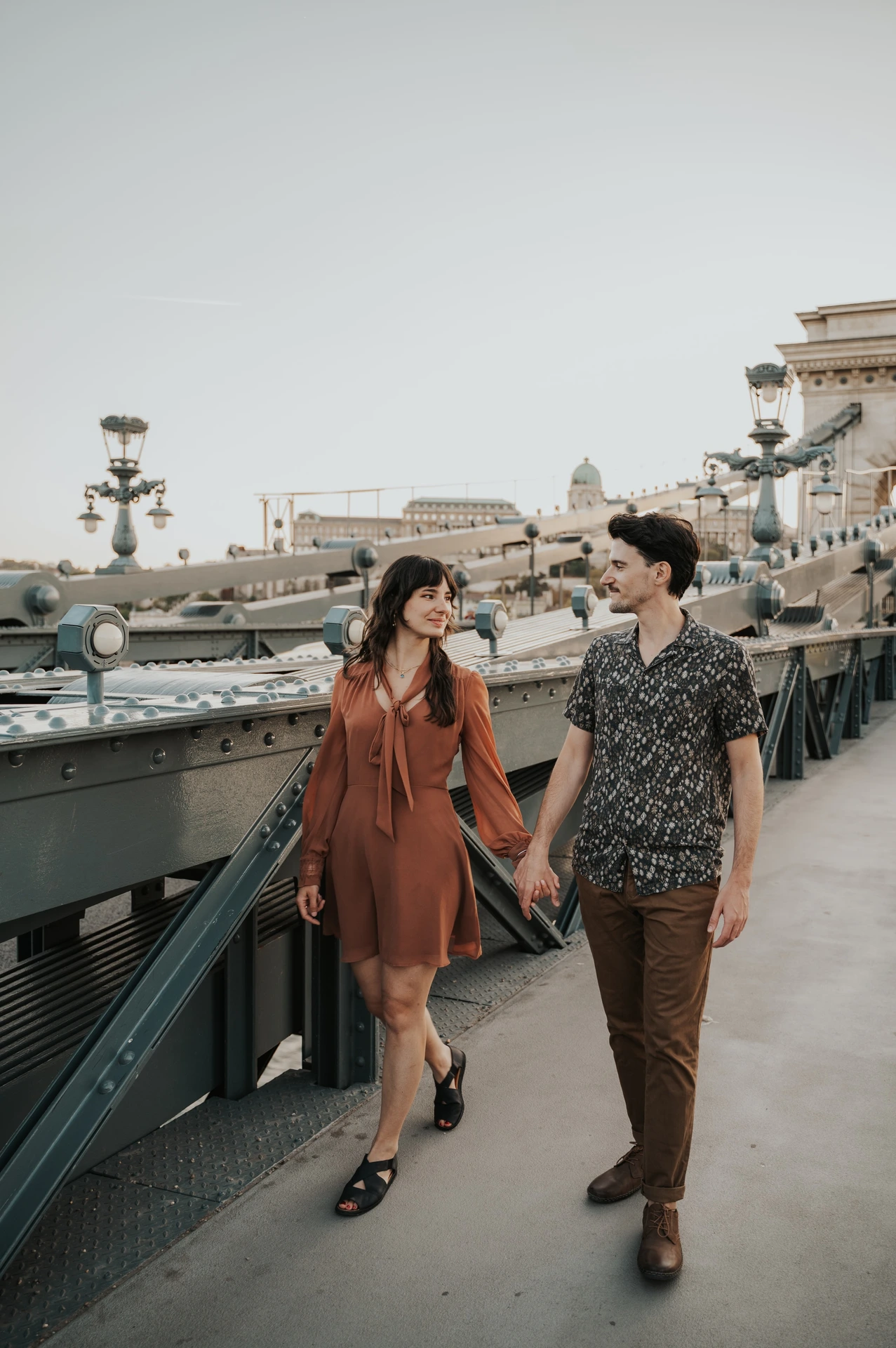 Budapest engagement photoshoot: A wide bridge walkway portrait A wide vertical view of the couple walking hand in hand along Chain Bridge, framed by its railings and street lamps during their Budapest engagement photoshoot.