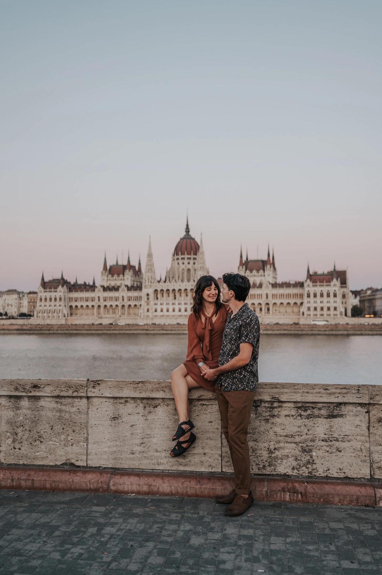 A wide vertical view of the couple kissing by the riverside stone wall, with Parliament filling the view across the water during their Budapest engagement photoshoot.