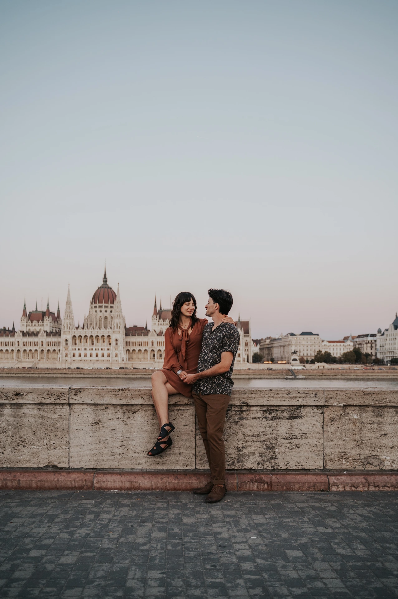 A wide vertical view of the couple sharing a kiss by the river wall, with Parliament’s dome and façade across the water during their Budapest engagement photoshoot.