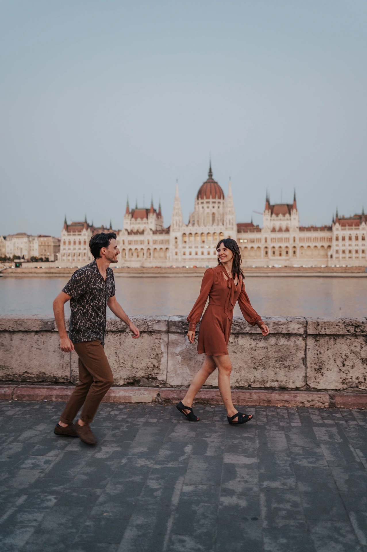 A wide vertical view of the couple holding hands during their riverside walk, with Parliament’s dome above the stone wall during their Budapest engagement photoshoot.