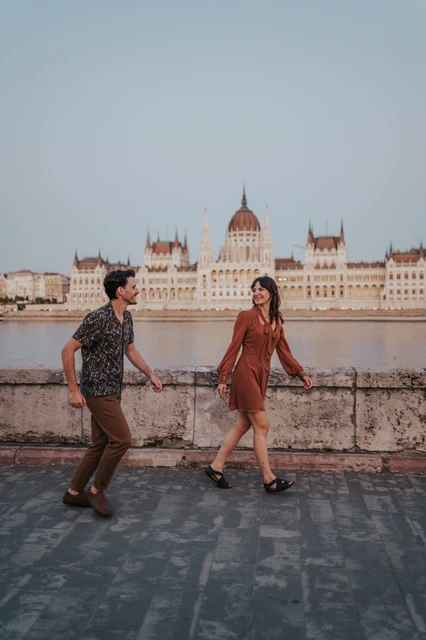Budapest engagement photoshoot: A wide portrait walking beside the river wall A wide vertical view of the couple holding hands during their riverside walk, with Parliament’s dome above the stone wall during their Budapest engagement photoshoot.
