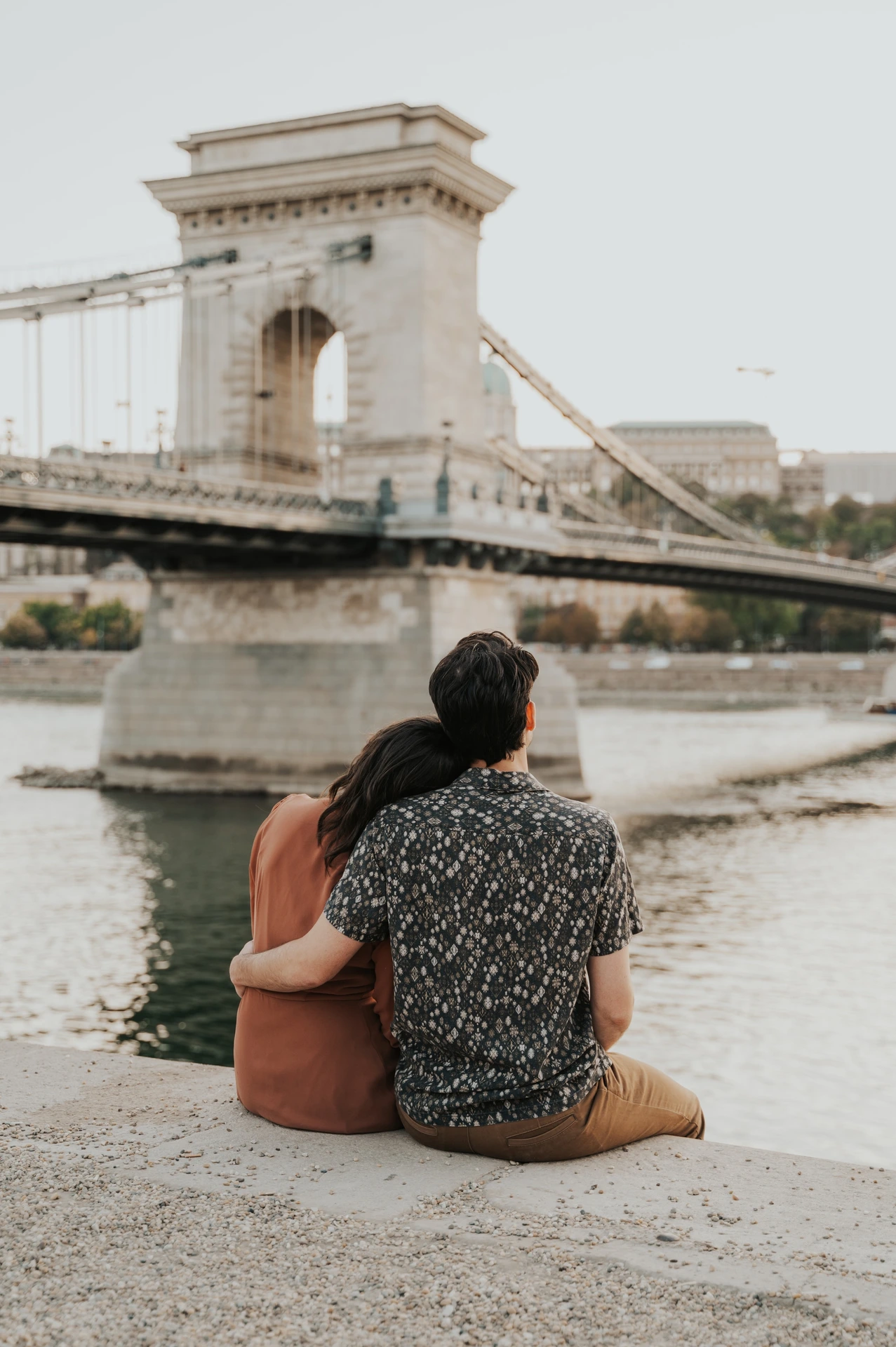 Budapest engagement photoshoot: A wider seated view across the river A wider vertical photograph of the couple sitting together by the river, with Chain Bridge’s tower and suspension chains above them during their Budapest engagement photoshoot.