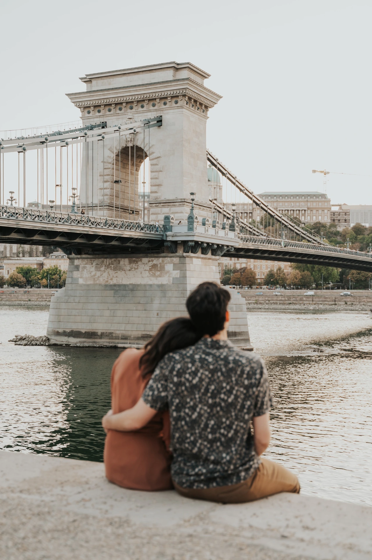 Budapest engagement photoshoot: An arm around a shoulder by the Danube The couple seated beside the Danube with an arm around a shoulder, facing Chain Bridge across the water during their Budapest engagement photoshoot.