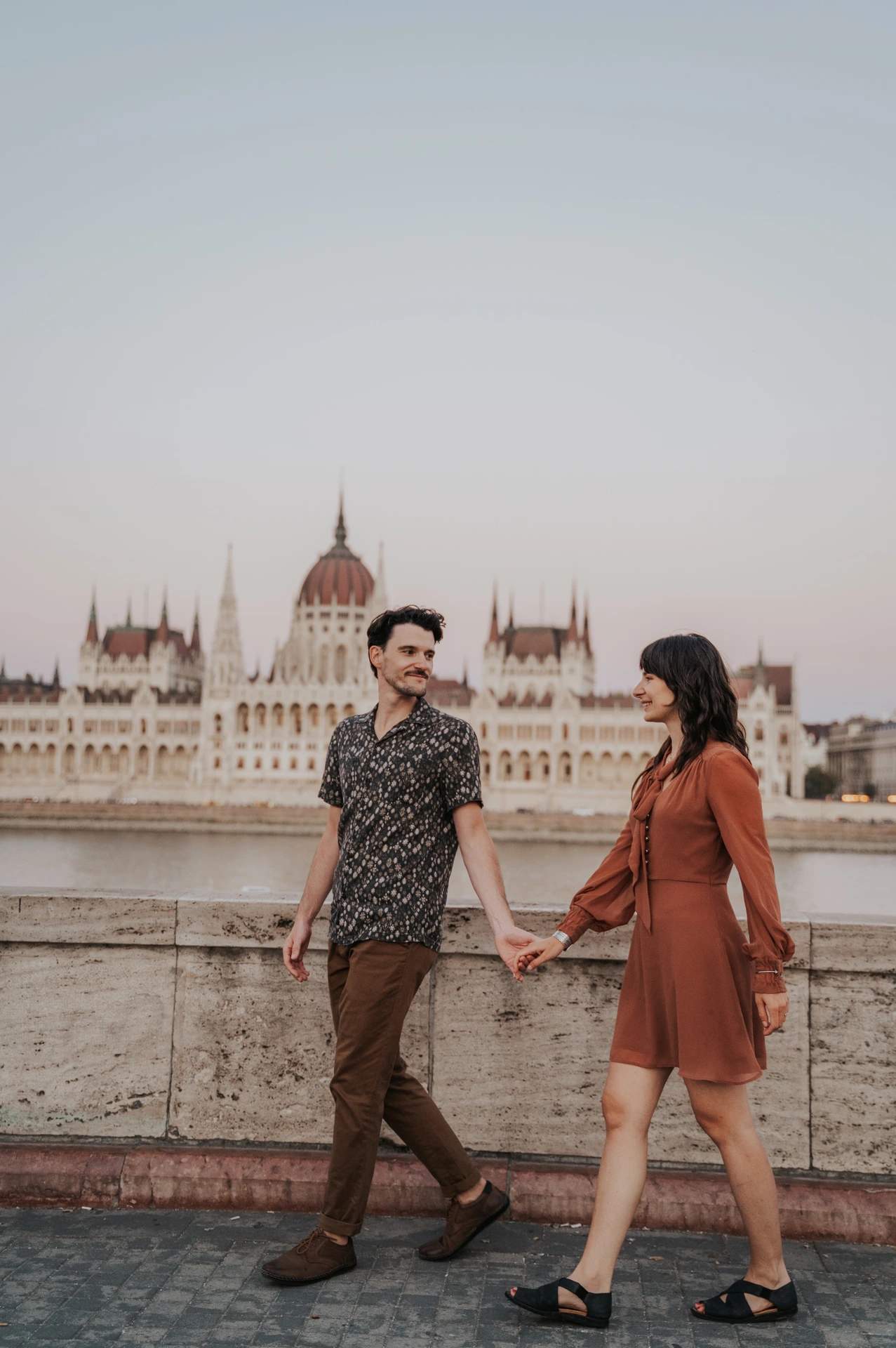 Budapest engagement photoshoot: An easy walk along the riverside wall The couple holding hands and walking along the riverside paving, with Parliament and the Danube in the background during their Budapest engagement photoshoot.