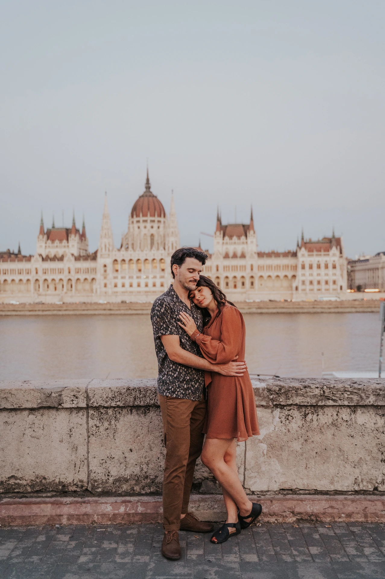 The couple standing in an embrace beside the river wall, with Parliament’s dome and pointed towers across the Danube during their Budapest engagement photoshoot.