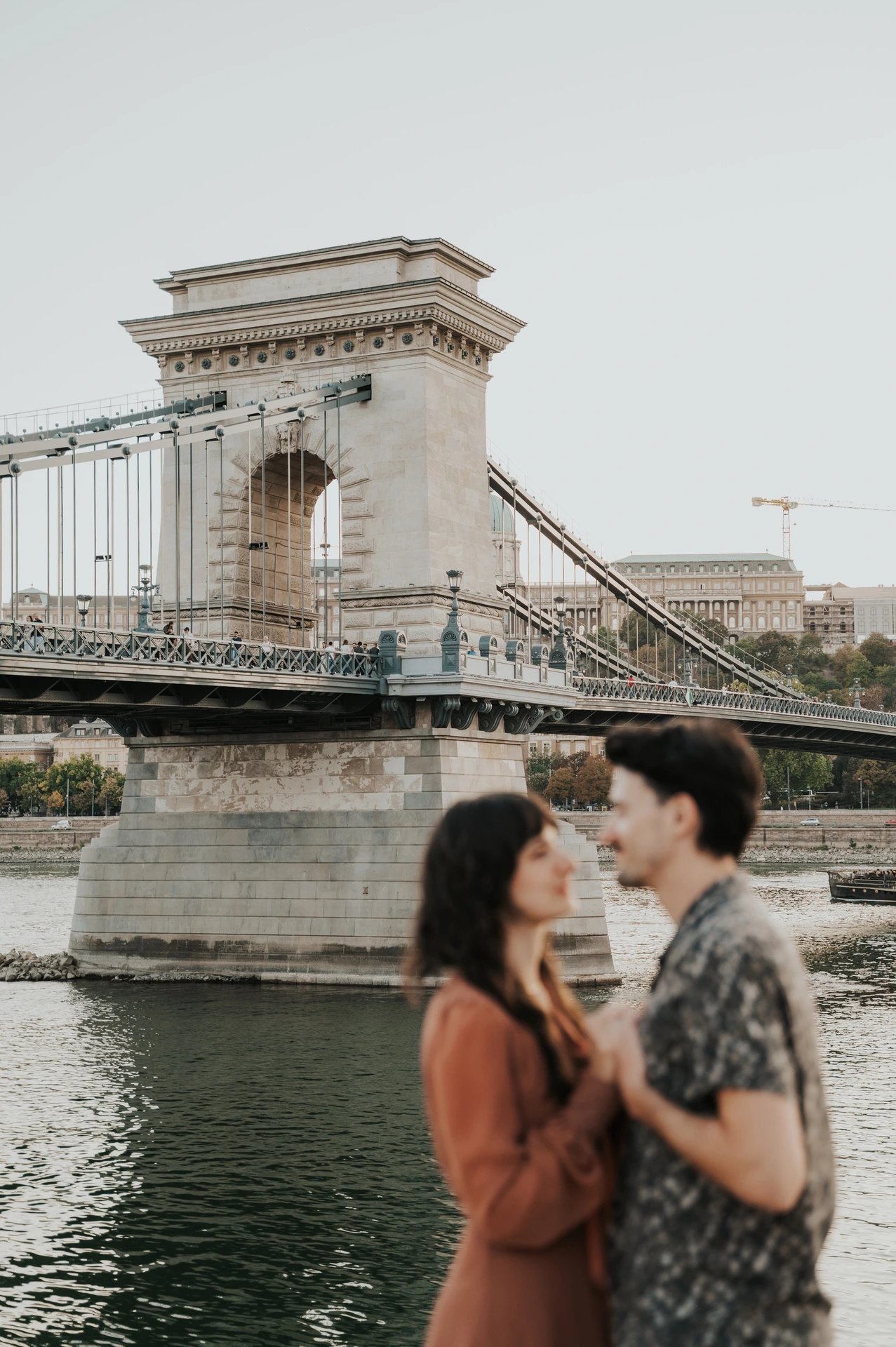 Budapest engagement photoshoot: An embrace framed by the bridge tower The couple standing close and holding hands by the river, with Chain Bridge’s tower and suspension chains in the background during their Budapest engagement photoshoot.