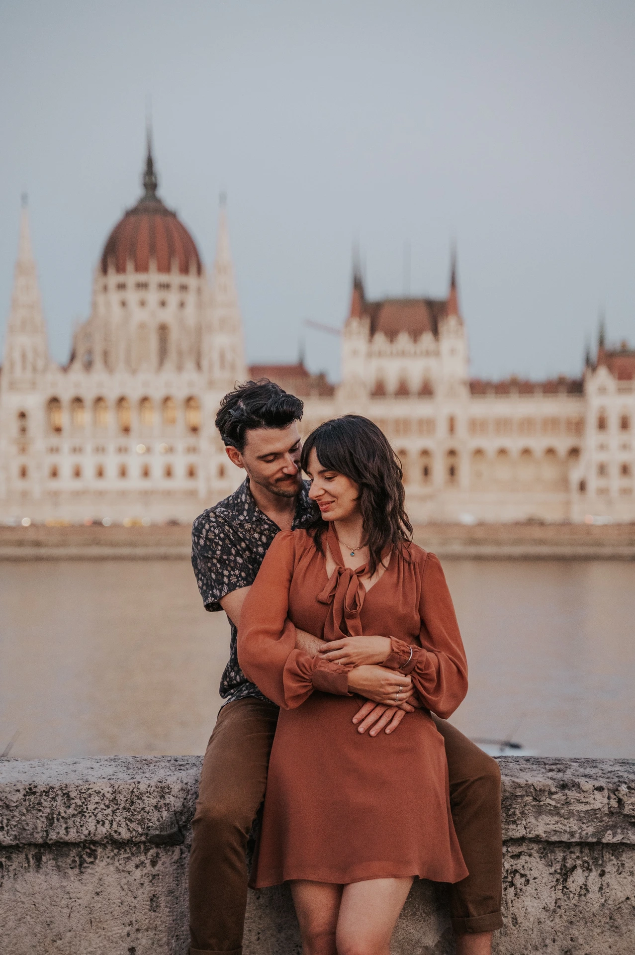 Budapest engagement photoshoot: An embrace from behind opposite Parliament The couple embracing from behind and looking down with gentle smiles, with Parliament softly blurred across the river during their Budapest engagement photoshoot.